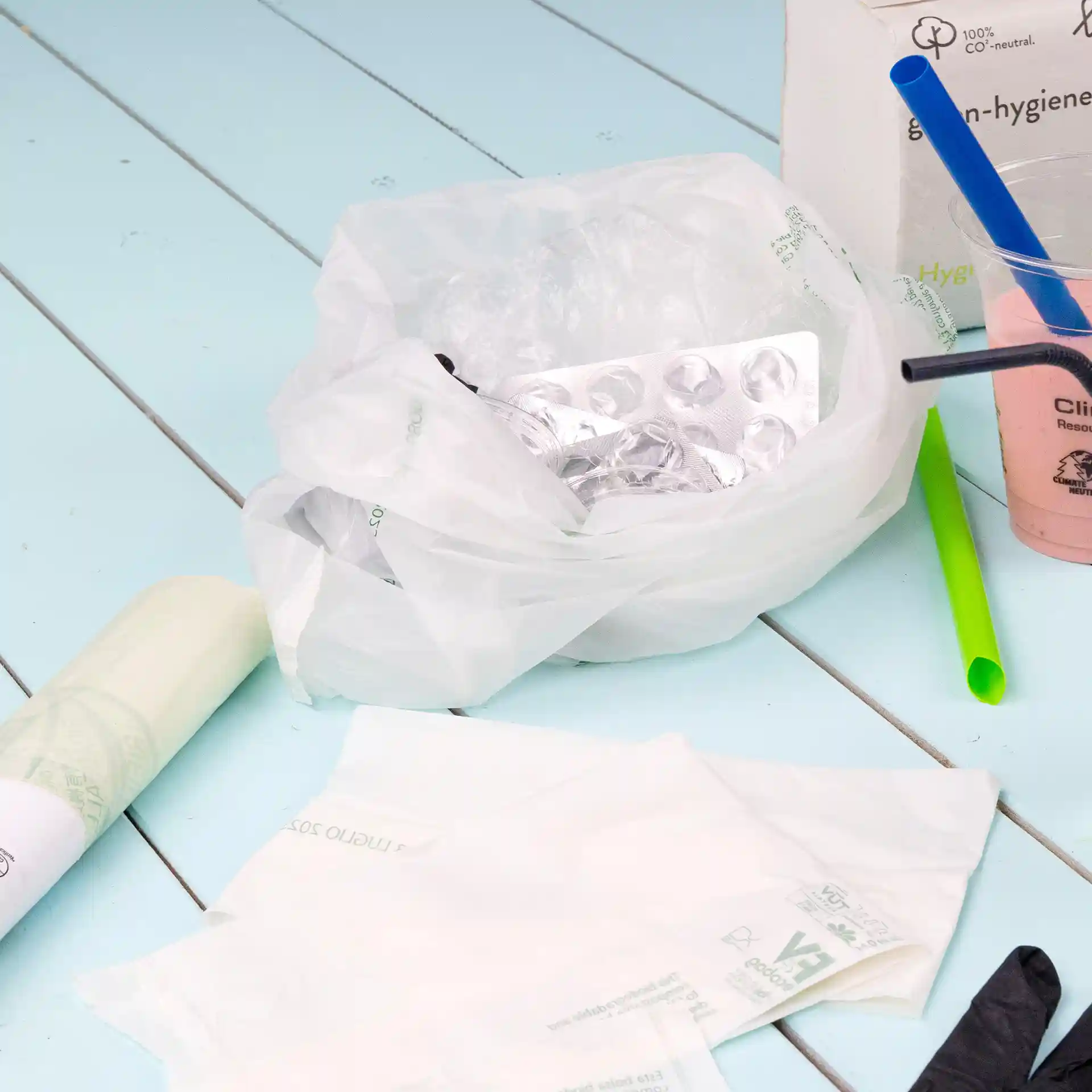 Sacchetto bio trasparente con contenuto tra cui un blister, cannucce di plastica e articoli usa e getta su una superficie blu.