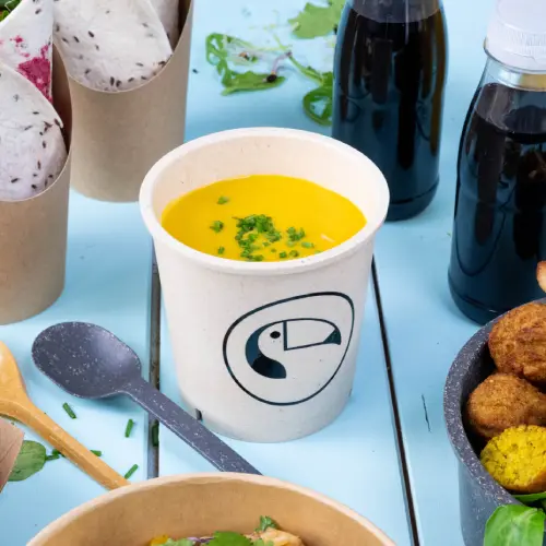 Tazza di zuppa con zuppa gialla e decorazioni verdi, circondata da contenitori per alimenti e bevande su una superficie blu.