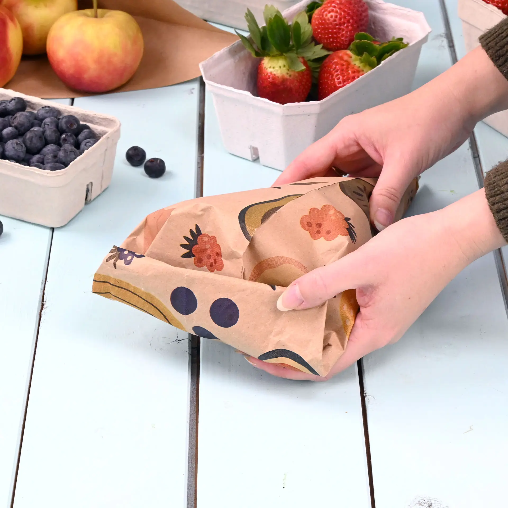 Mani che tengono un cono di carta stampato personalizzato con un design marrone, circondato da cesti di frutta su un tavolo di legno.
