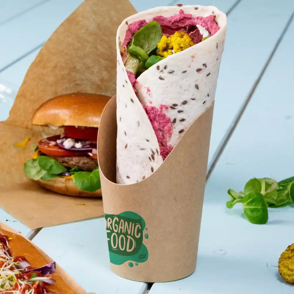 Wrap ripieno di ingredienti colorati in un contenitore di carta marrone, accanto a un hamburger e insalata su una superficie azzurra.