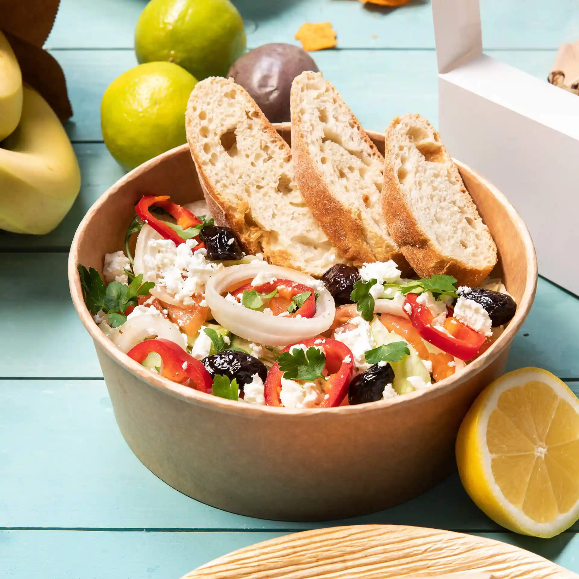 Ciotola di carta rotonda marrone piena di insalata, guarnita con pane affettato, circondata da frutta e una scatola da asporto bianca.
