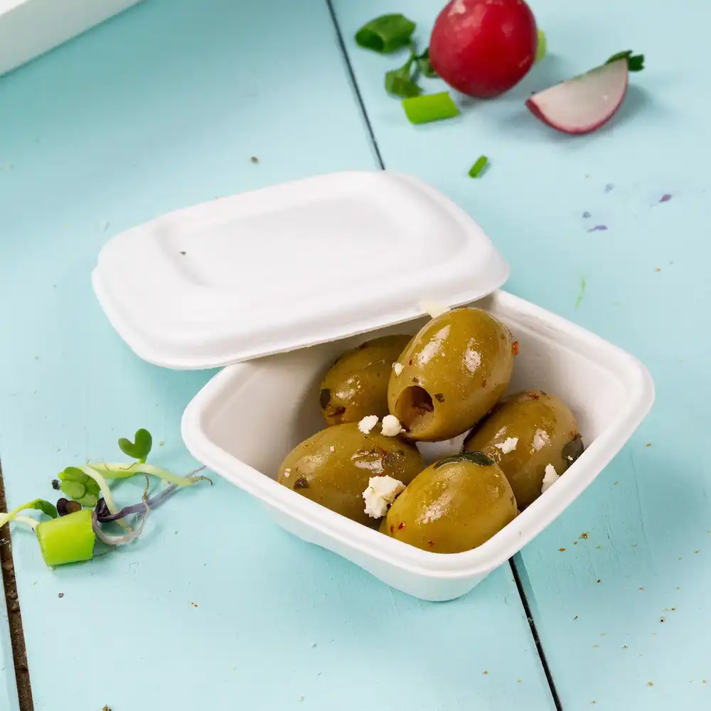 Contenitore con diverse olive verdi con condimenti, su uno sfondo azzurro, con una rapa e erbe vicine.