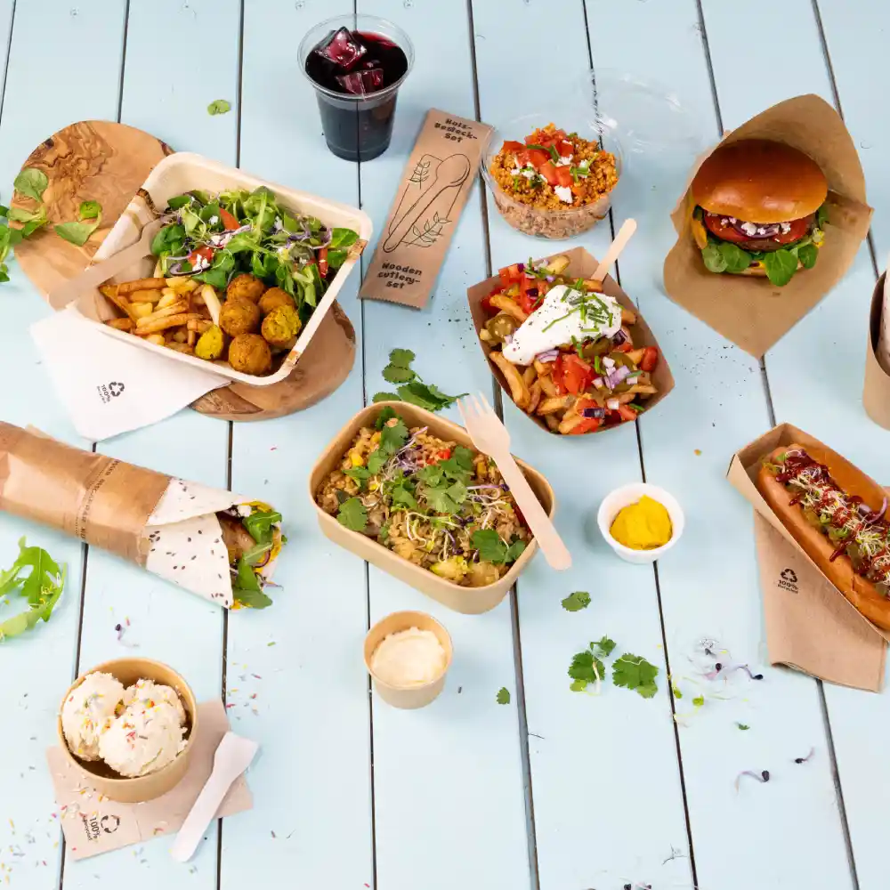 Un layout di vari piatti da catering su un tavolo azzurro chiaro, comprese insalate, hamburger, hot dog e dolci. È inclusa anche una bevanda.