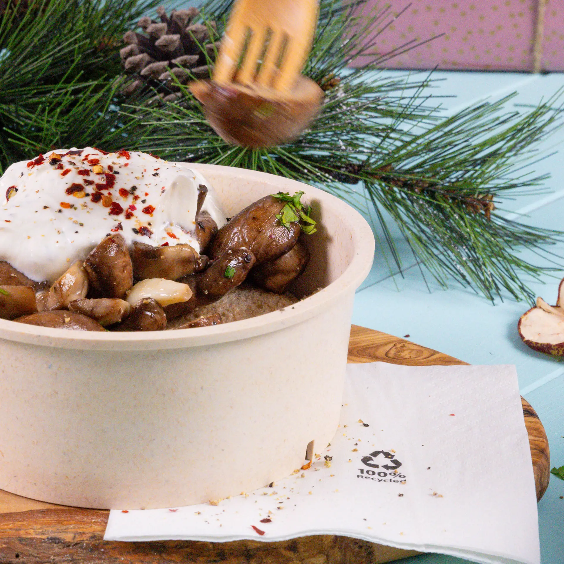 Piatto appena preparato in una ciotola di cartone marrone, guarnito con yogurt, erbe e spezie; accompagnato da un tovagliolo da cocktail bianco riciclato.