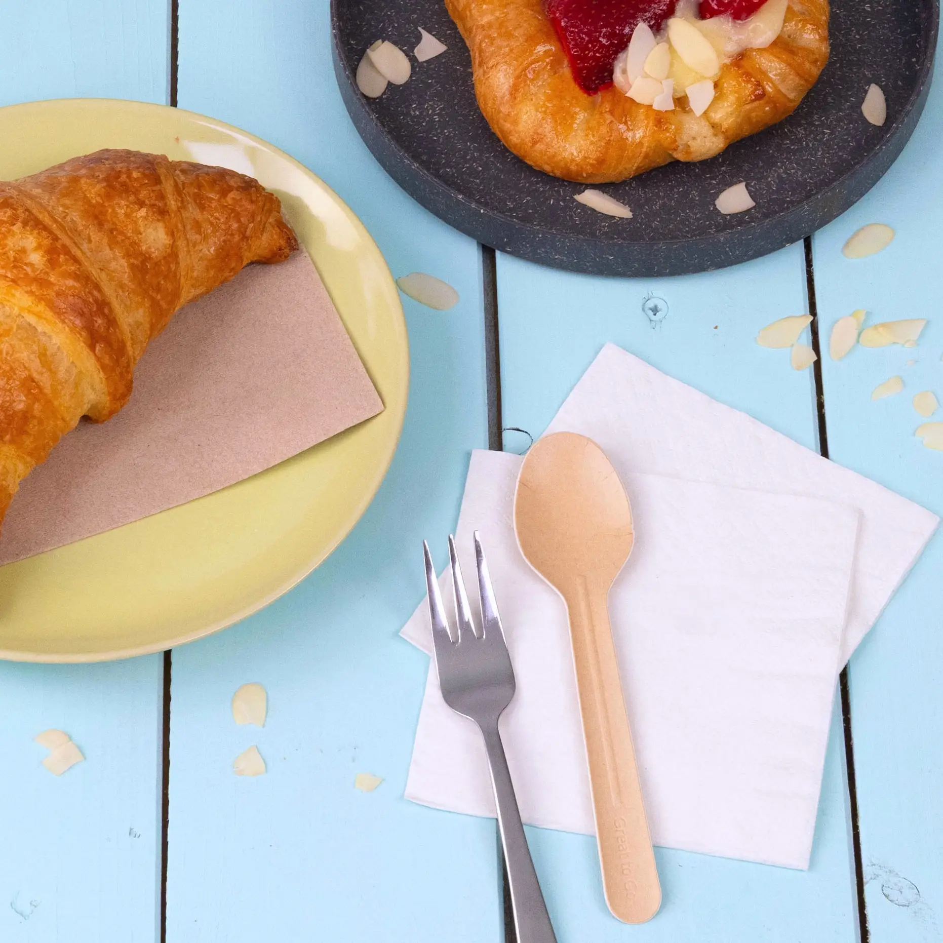 Immagine di un croissant su un piatto giallo, un dolce su un piatto scuro, un cucchiaio di carta, una forchetta e tovaglioli su un tavolo di legno blu.