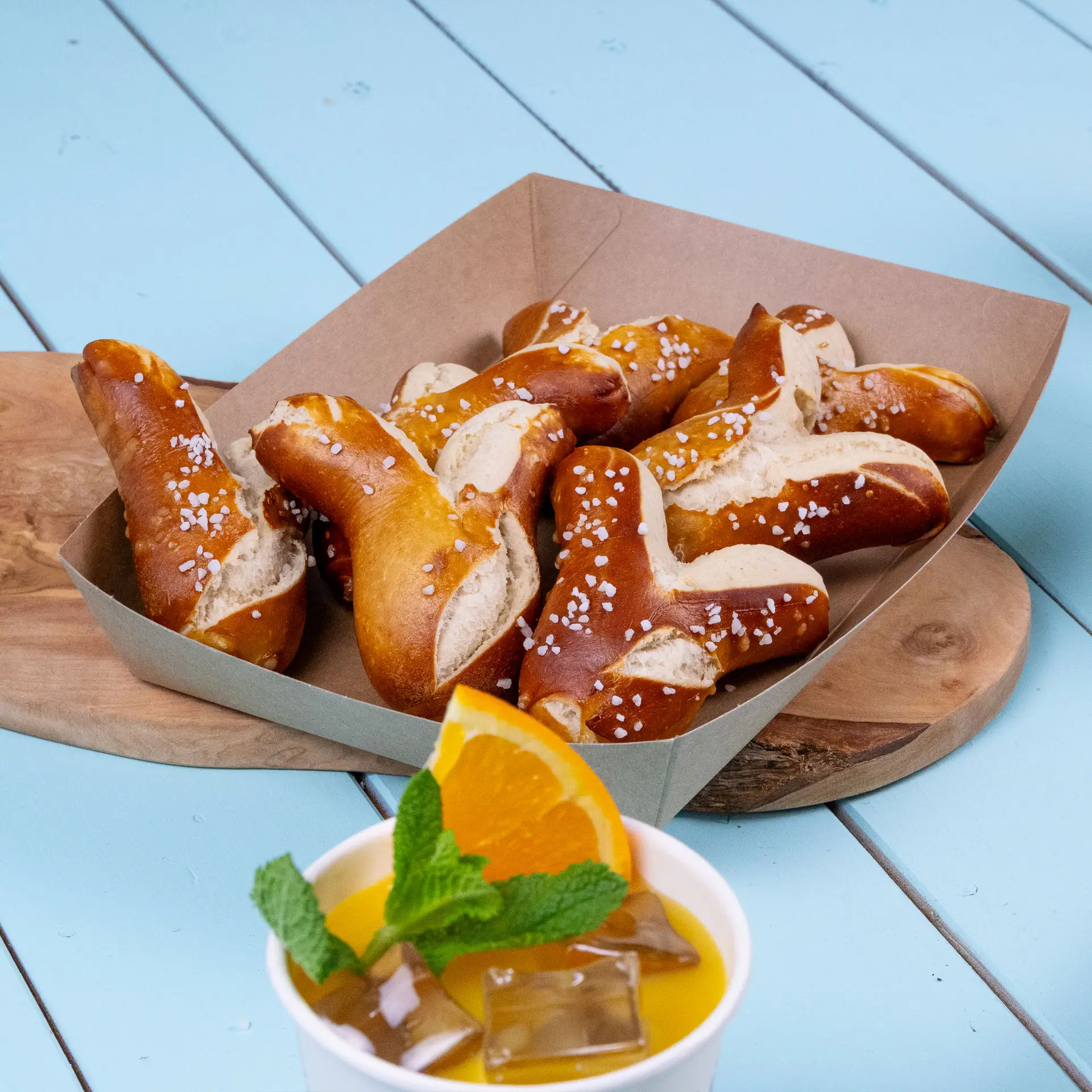 Vassoio di cartone marrone riempito di pretzel guarniti con sale grosso, accanto a una tazza di bevanda all'arancia decorata con foglie di menta.