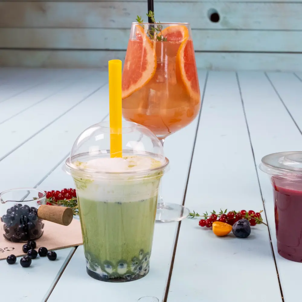 Una bevanda verde con una cannuccia gialla in un bicchiere trasparente, con perle di tapioca blu sul fondo; un cocktail colorato con fette di pompelmo in un bicchiere dietro.