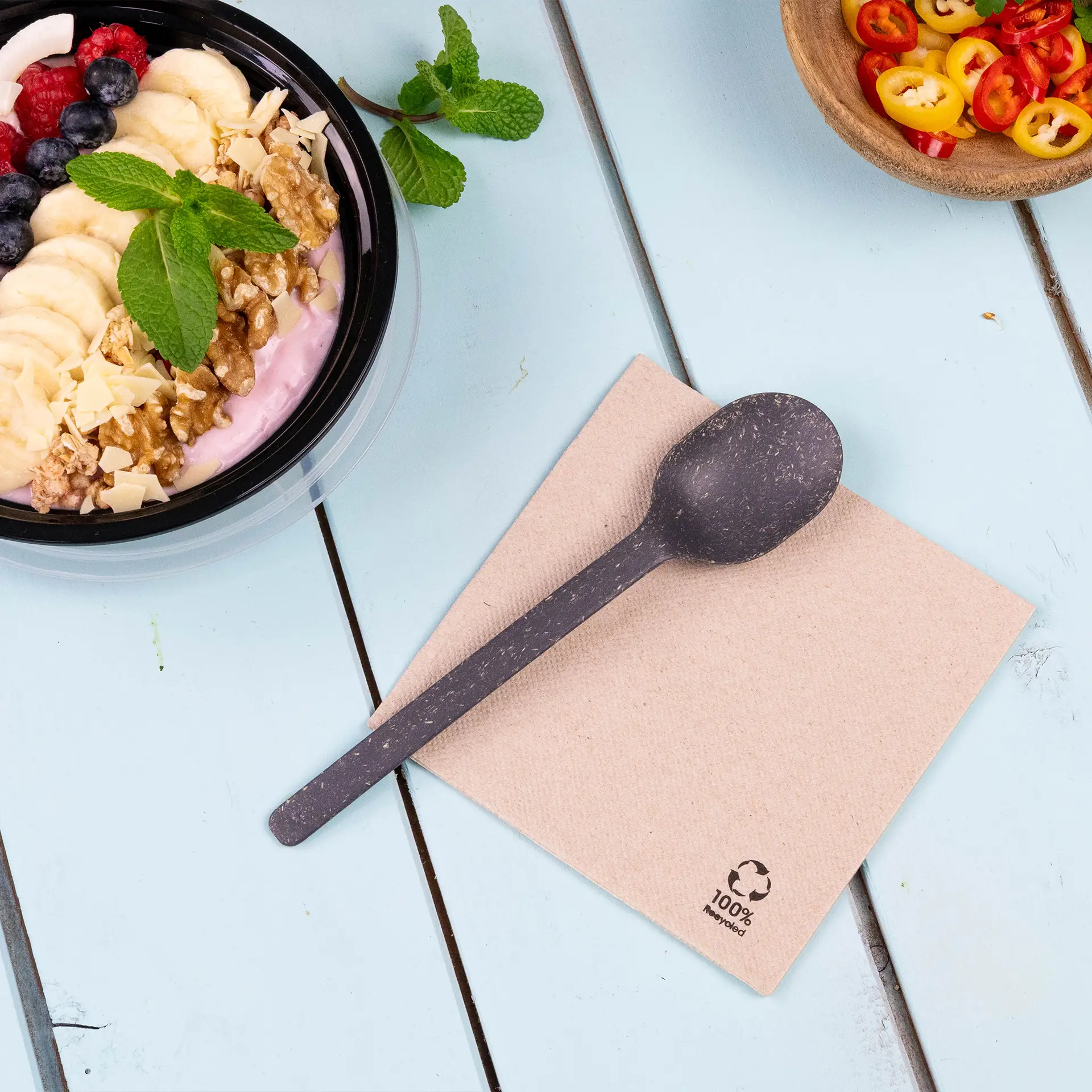 Tovagliolo da cocktail in carta riciclata con un cucchiaio scuro biodegradabile su una superficie azzurro chiaro accanto a una ciotola di yogurt con frutta.