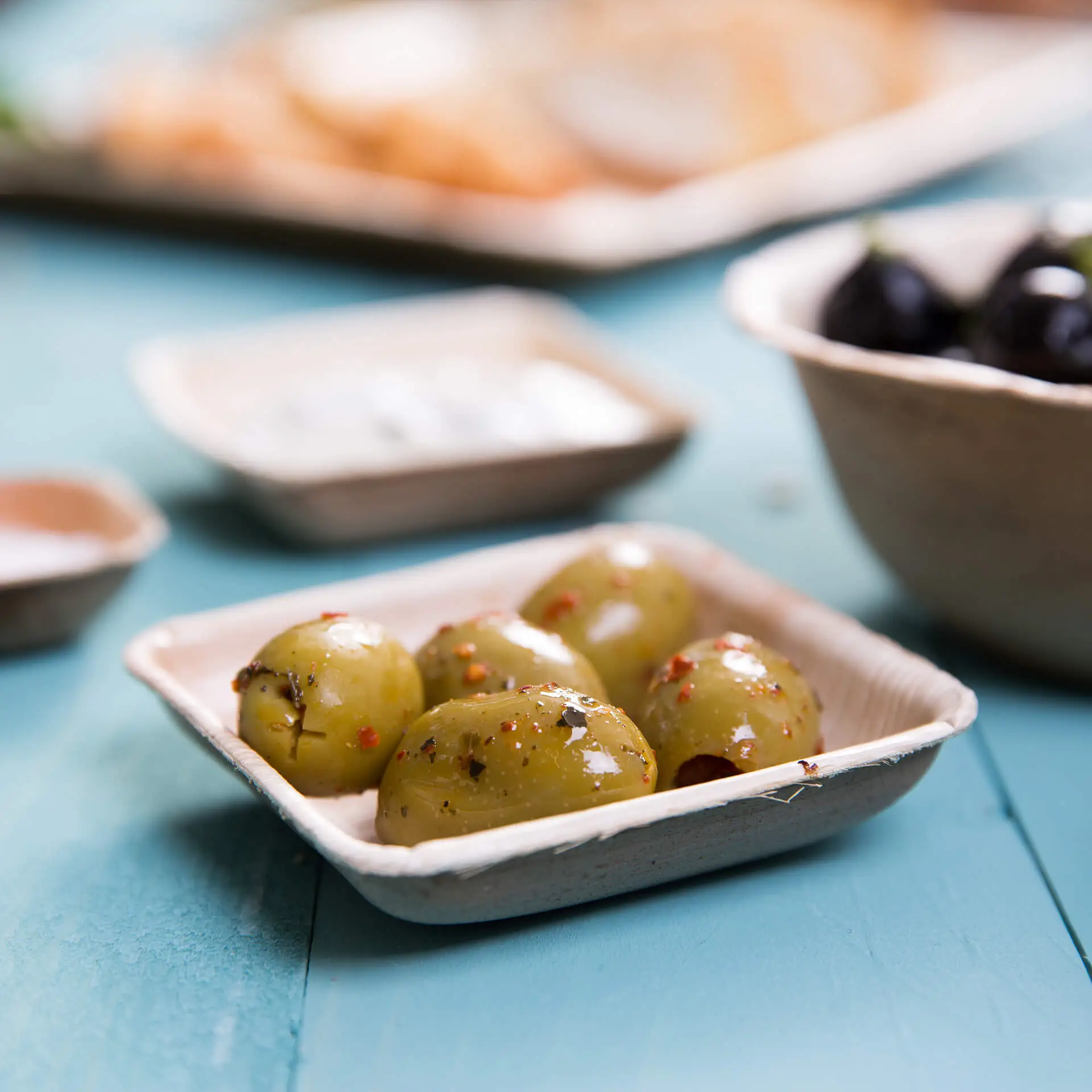 Ciotola quadrata in foglia di palma contenente diverse olive verdi con spezie; articolo per la tavola leggero ed ecologico.
