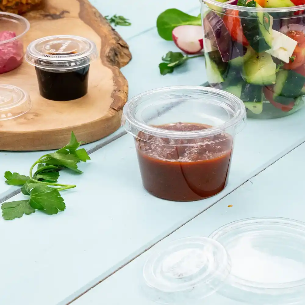 Contenitore trasparente con salsa rossastra, accanto a erbe verdi e altri alimenti su una superficie azzurro chiaro.