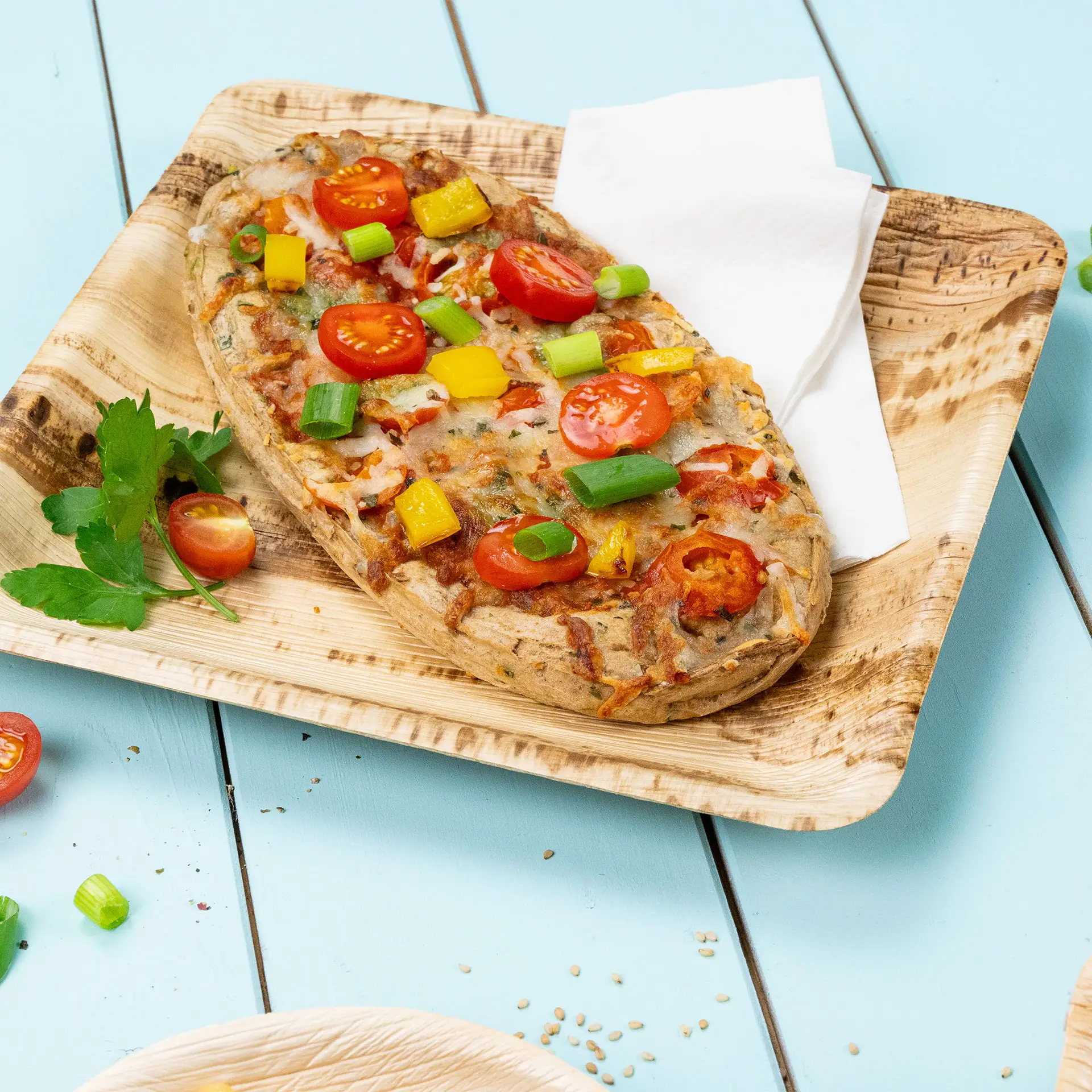 Un piatto quadrato in foglia di palma con pane piatto guarnito con pomodorini, peperoni e cipolle verdi su uno sfondo azzurro chiaro.