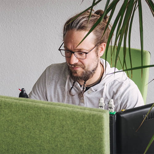 Uomo con occhiali che lavora a una scrivania, concentrato su un computer; piante e decorazioni d'ufficio verdi visibili sullo sfondo.