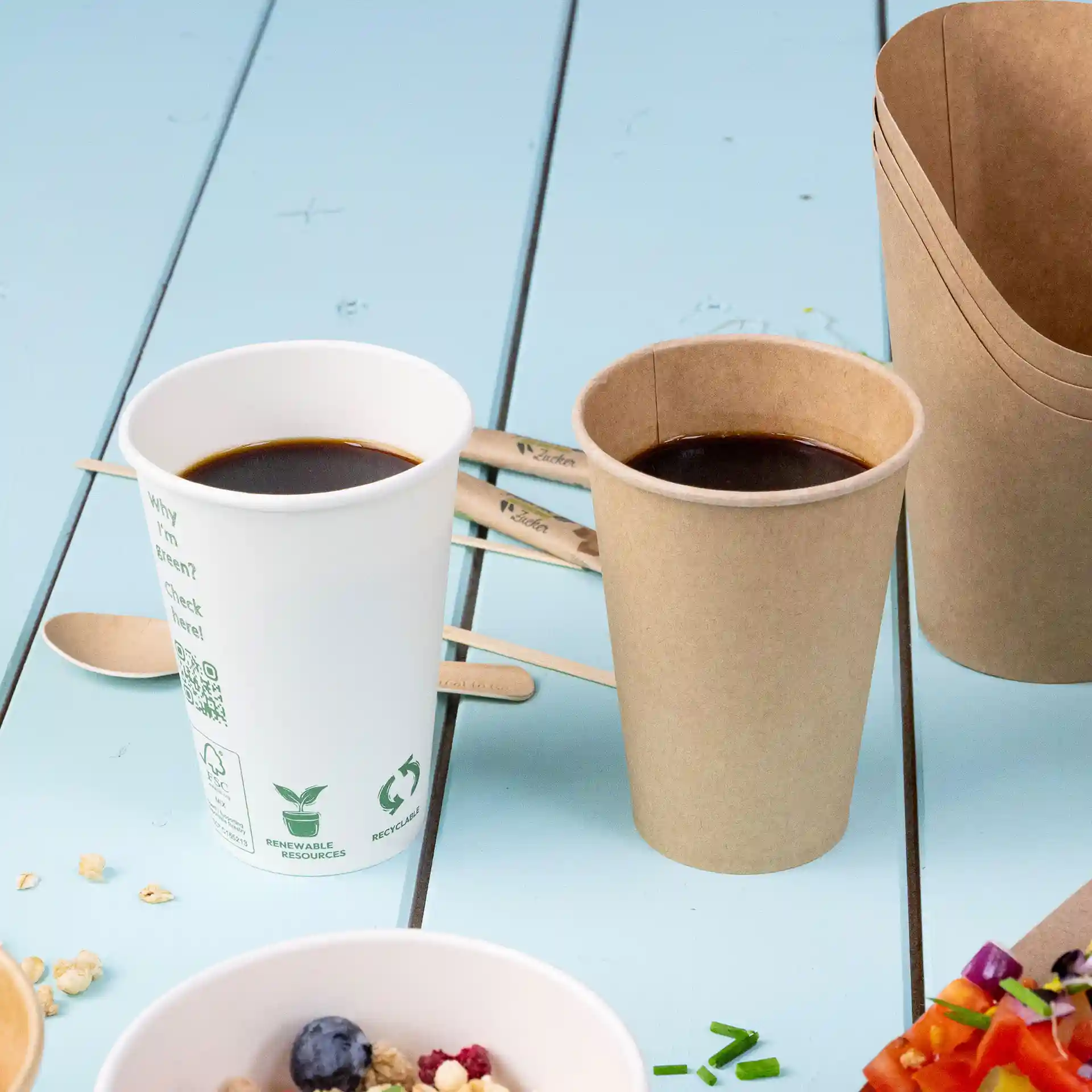 Bicchiere per distributore in PLA bianco con caffè, accanto a un bicchiere di carta marrone, su una superficie blu con cucchiai di legno e cibo.