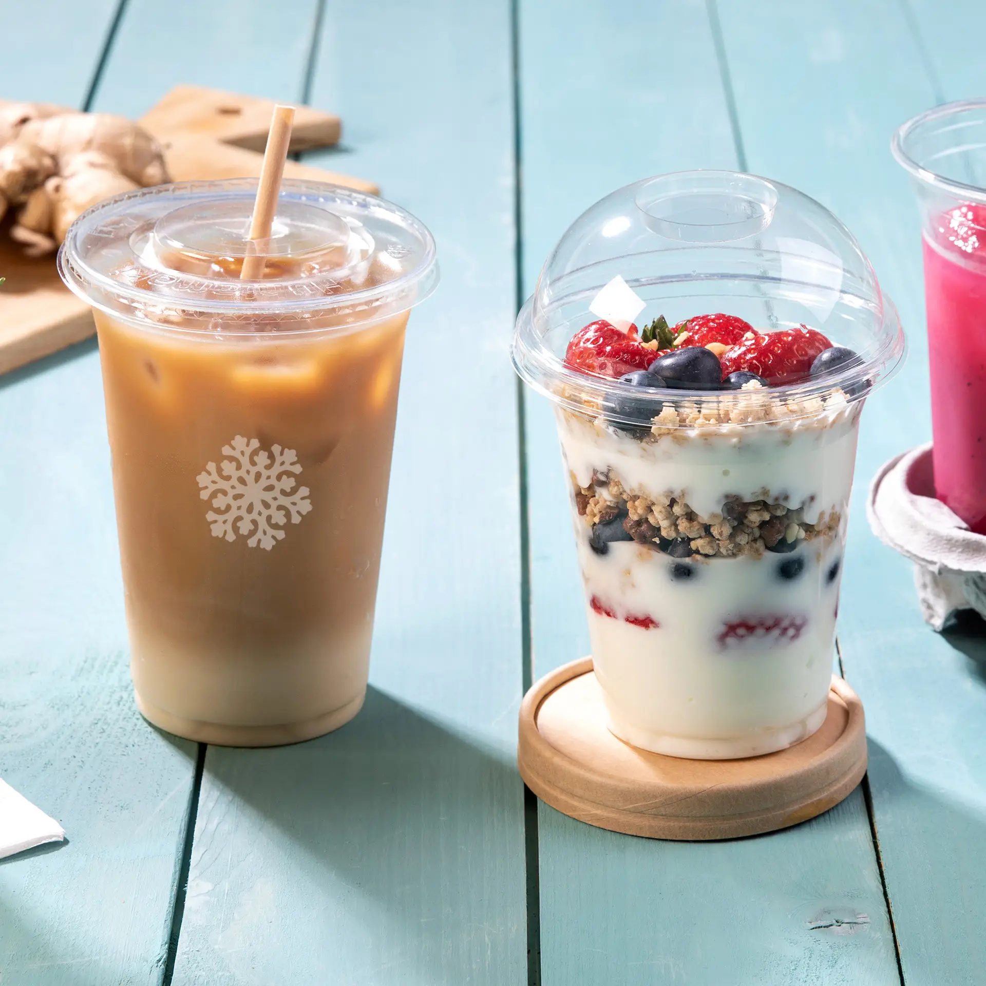 Tre bicchieri PLA trasparenti per bevande fredde: un bicchiere di caffè freddo, un parfait con frutti di bosco e una bevanda rosa, su un tavolo di legno.