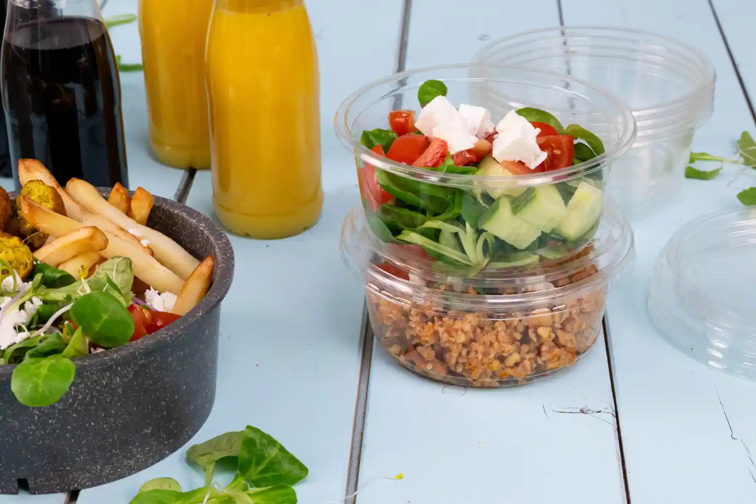 Contenitori con insalata e cereali accanto a una ciotola di patatine; due bottiglie di bevande sullo sfondo; superficie in legno blu chiaro.