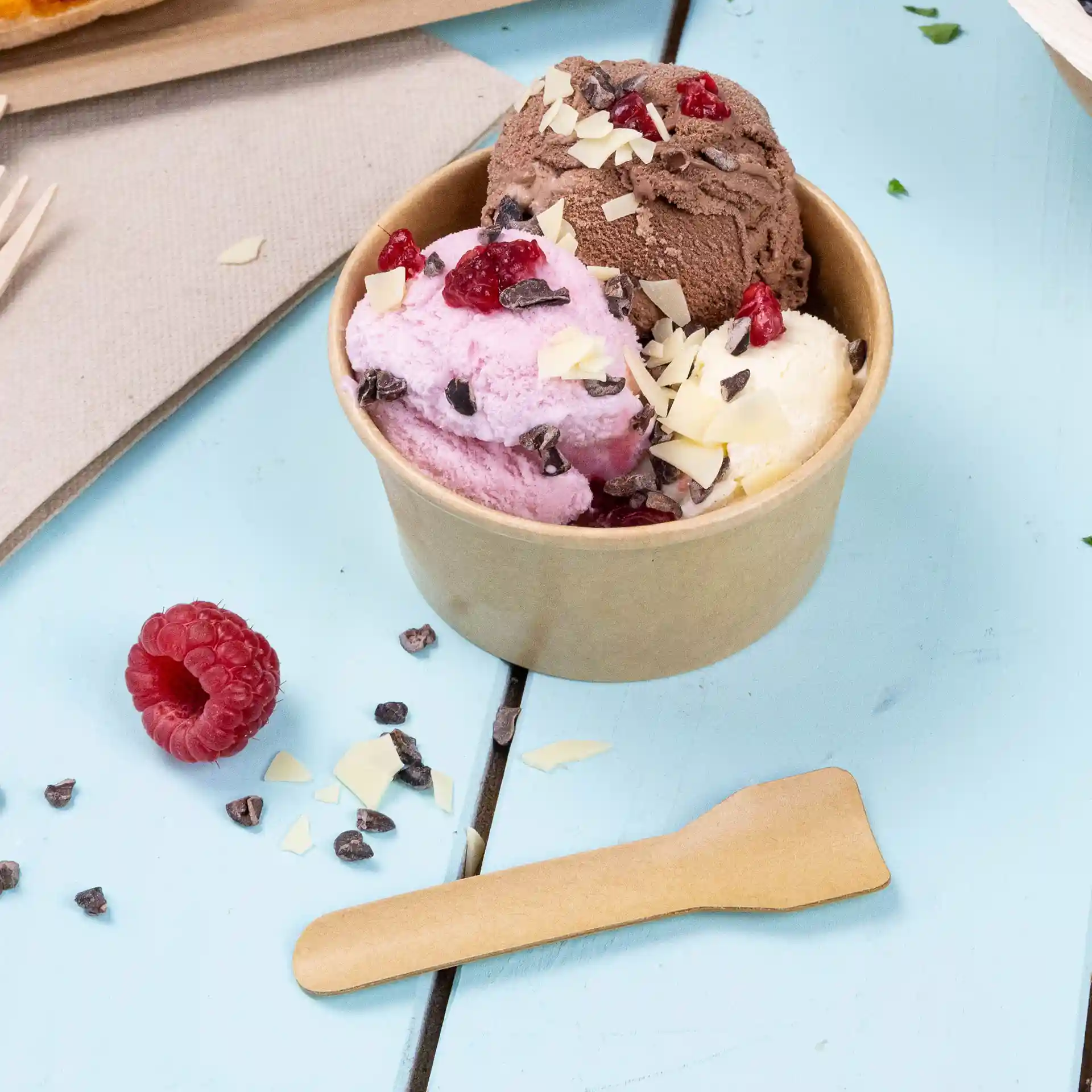 Tazza con tre palline di gelato rosa, marrone e bianco, guarnita con gocce di cioccolato e lamponi, con un cucchiaio di legno.