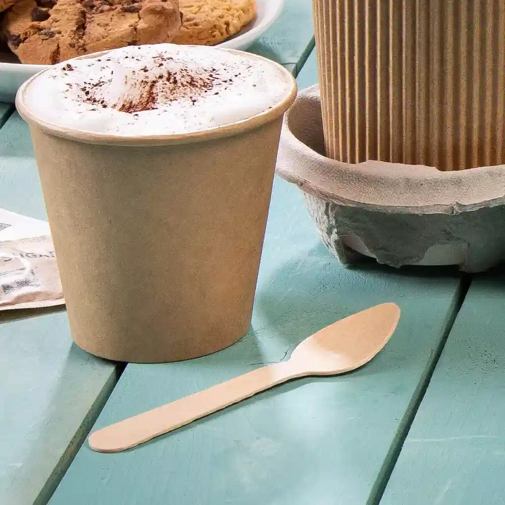 Bicchiere usa e getta marrone con sopra la schiuma e un cucchiaio di legno su un tavolo turchese, con biscotti sullo sfondo.