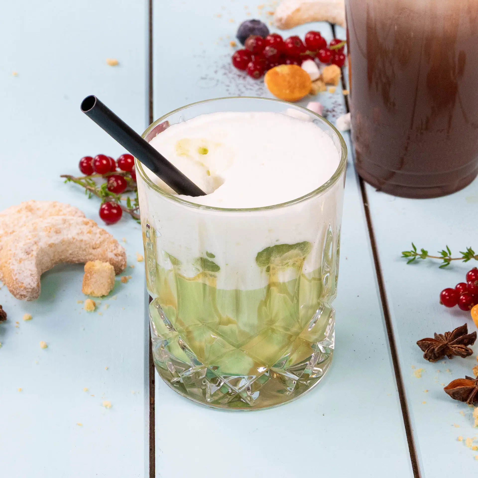 Un bicchiere riempito con bevanda verde a strati, guarnito con schiuma bianca e una cannuccia nera; circondato da biscotti e bacche.