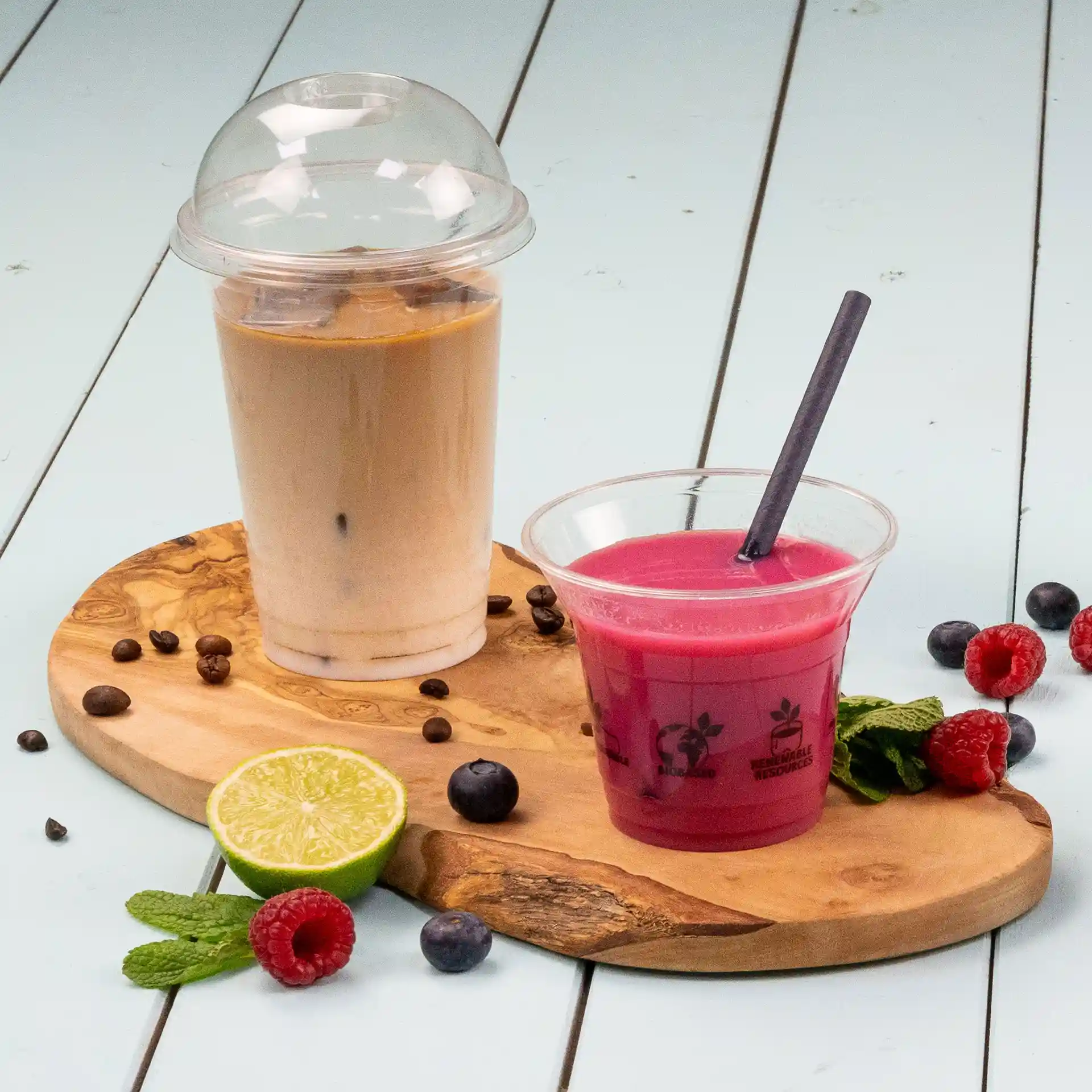 Immagine di un bicchiere trasparente con un coperchio a cupola riempito con una bevanda marrone chiaro, accanto a una bevanda rosa in un bicchiere trasparente con una cannuccia, su un tagliere di legno con frutta.