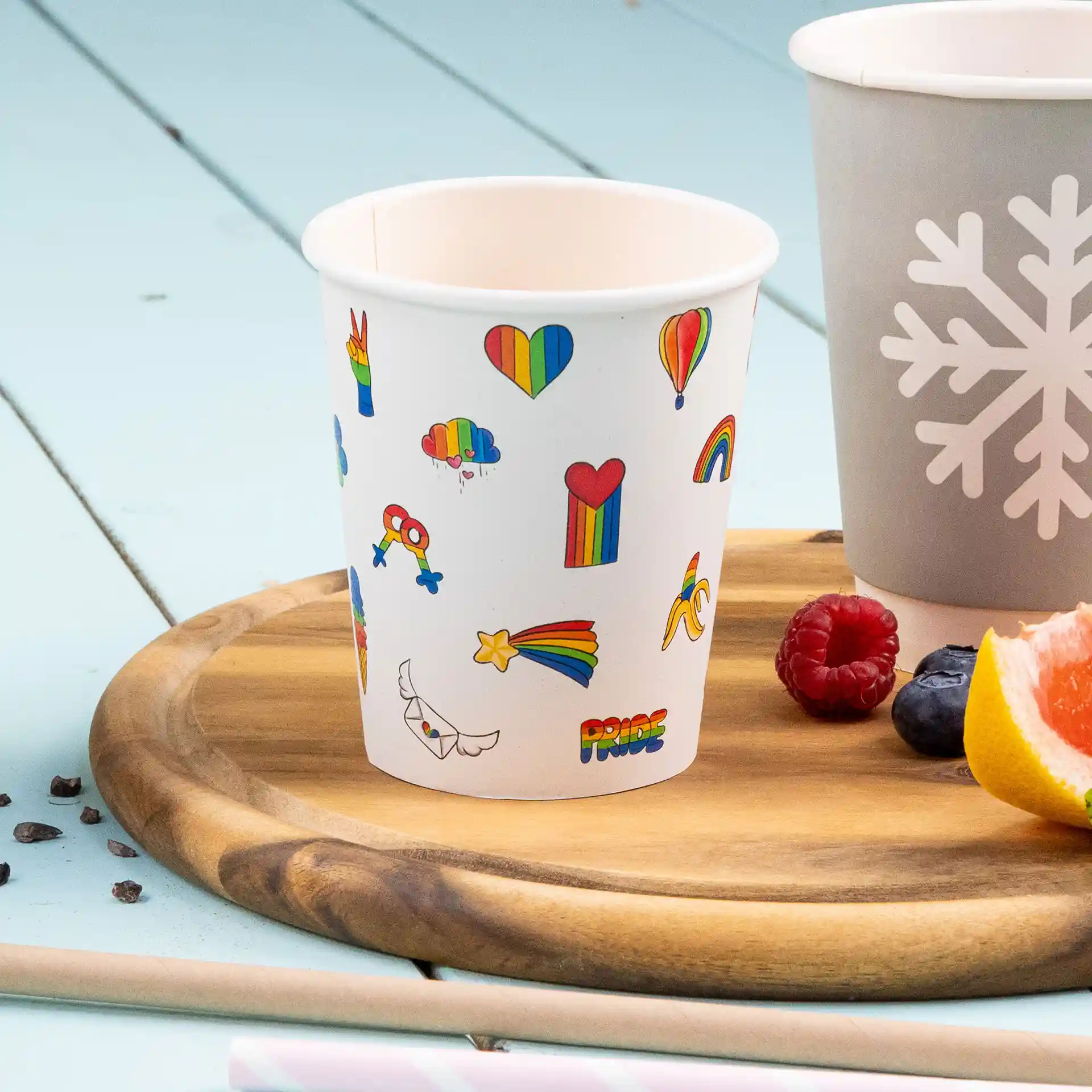 Bicchiere di carta bianco con design stampati colorati, tra cui cuori, arcobaleni e nuvole, su un vassoio di legno.