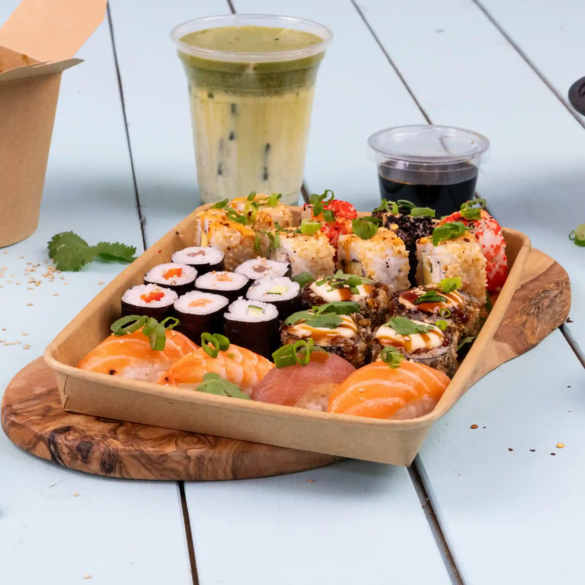 Vassoio di sushi assortiti, inclusi gamberi e salmone, con una bevanda e salsa di accompagnamento su un tagliere di legno.