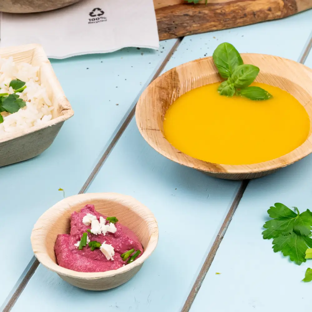 Ciotola in bambù riempita di zuppa gialla guarnita con una foglia di basilico; ciotola più piccola con salsa rosa guarnita con erbe; riso alle erbe in un contenitore vicino.