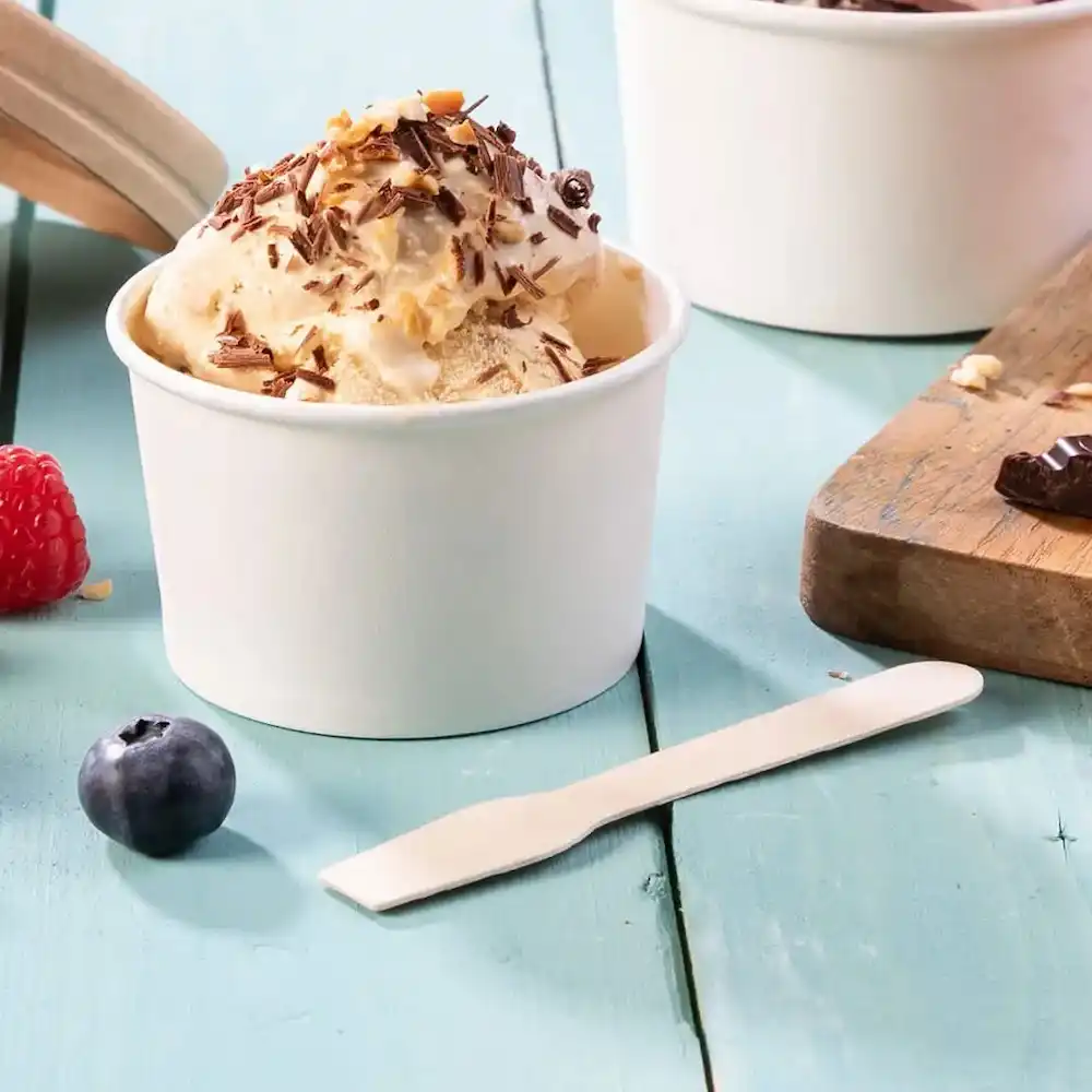 Cucchiaio usa e getta bianco accanto a un bicchiere di gelato guarnito con cioccolato e noci, con bacche su una superficie in legno blu.