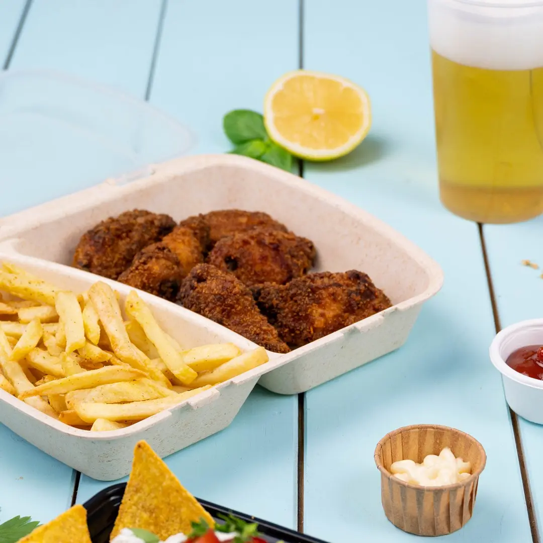 Contenitore da asporto con pollo fritto e patatine, fetta di limone, birra e salse per intingere su una superficie di legno blu.