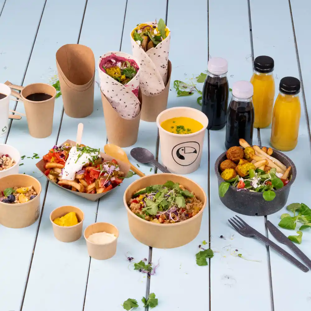 Varietà di alimenti in confezionamento ecologico, inclusi wrap, insalate, zuppe e bevande su una superficie di legno blu.