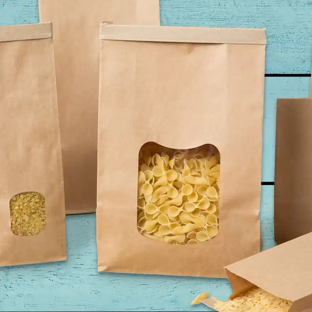 Sacchetti di carta marrone con finestre trasparenti, pieni di diverse forme di pasta, su una superficie di legno blu.