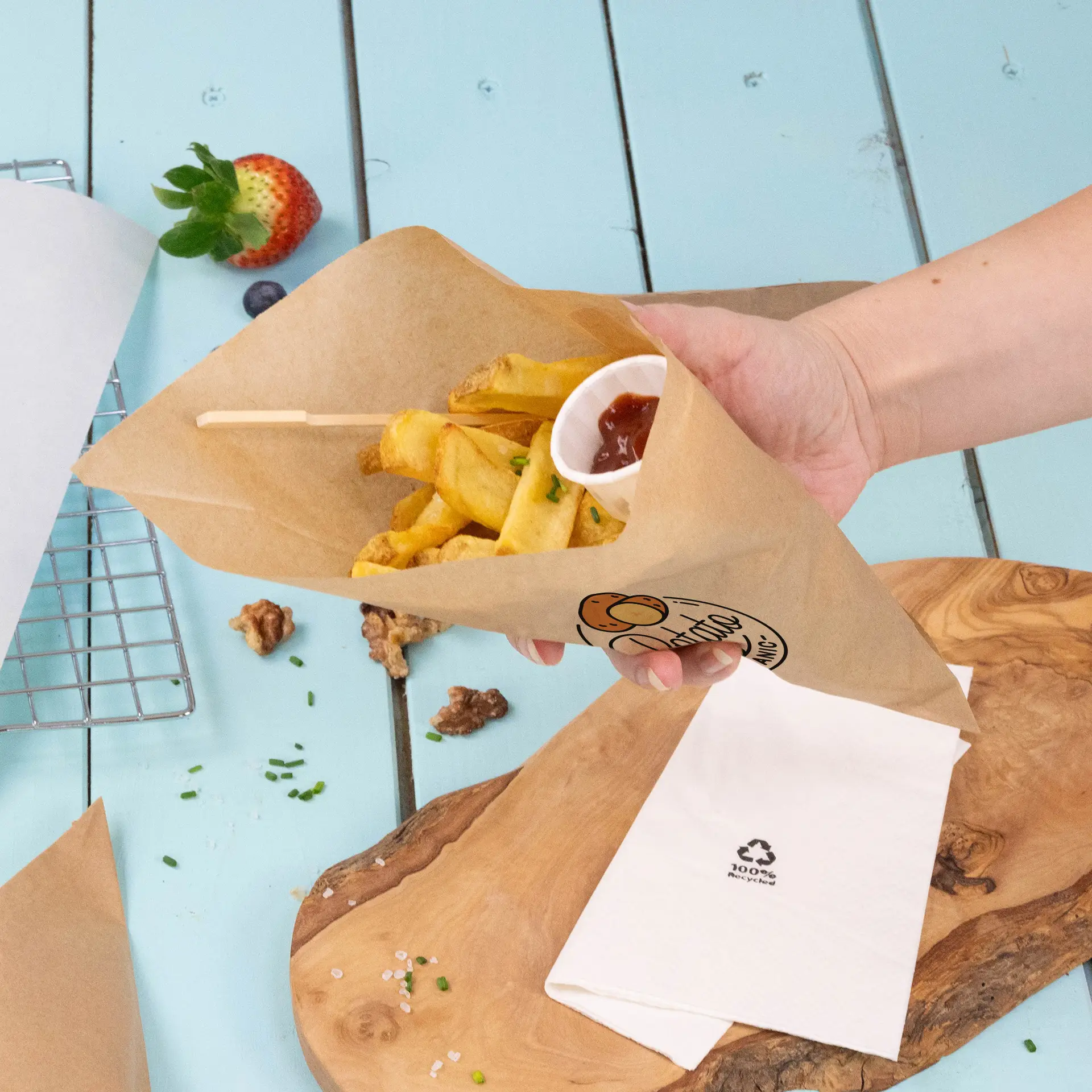 Mano che tiene un cono di carta marrone pieno di patatine e un piccolo bicchiere di ketchup, su una superficie di legno blu.
