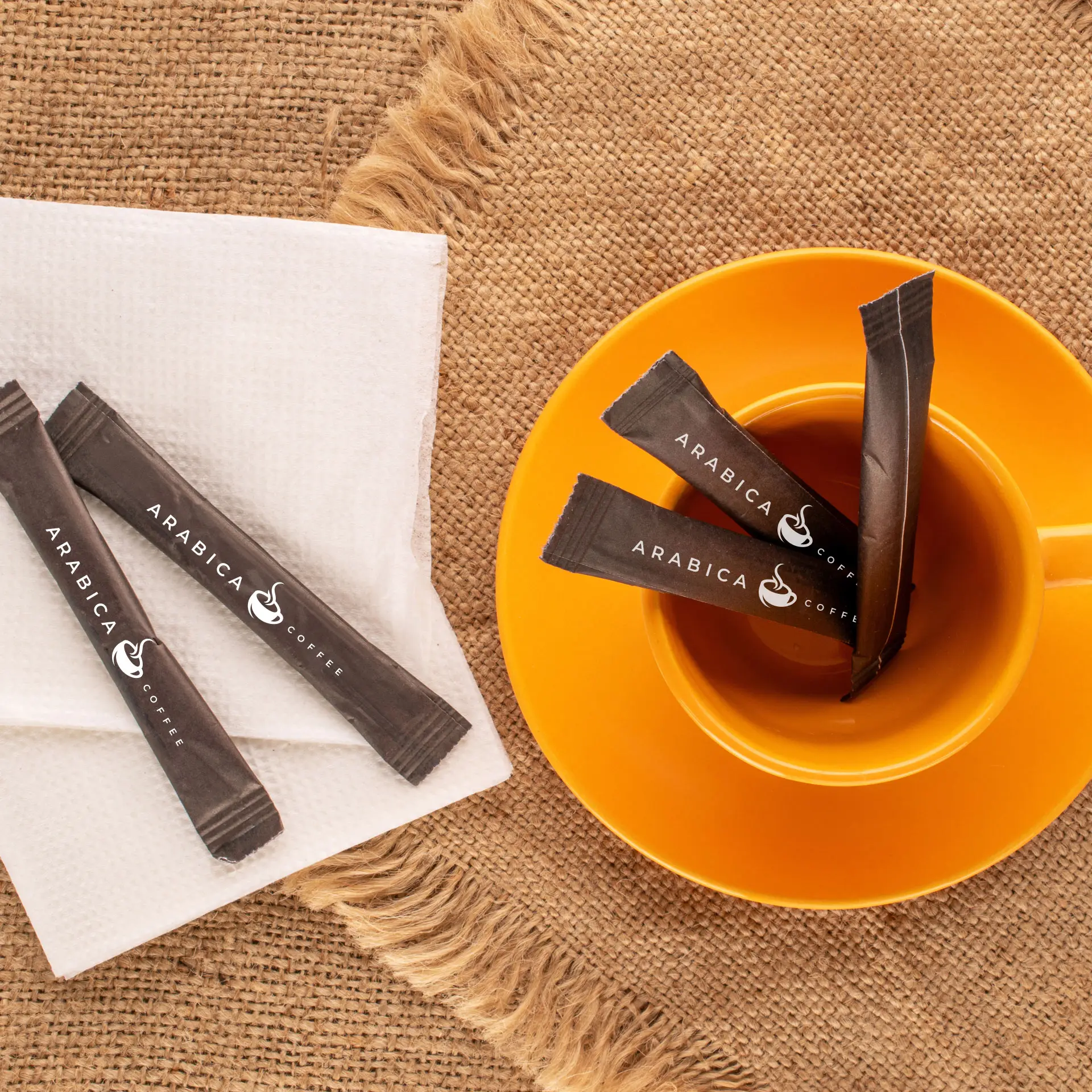 Diverse bastoncini di zucchero personalizzati con logo "Arabica Coffee" sono posizionati in una tazza arancione, accanto a un tovagliolo su una superficie testurizzata.