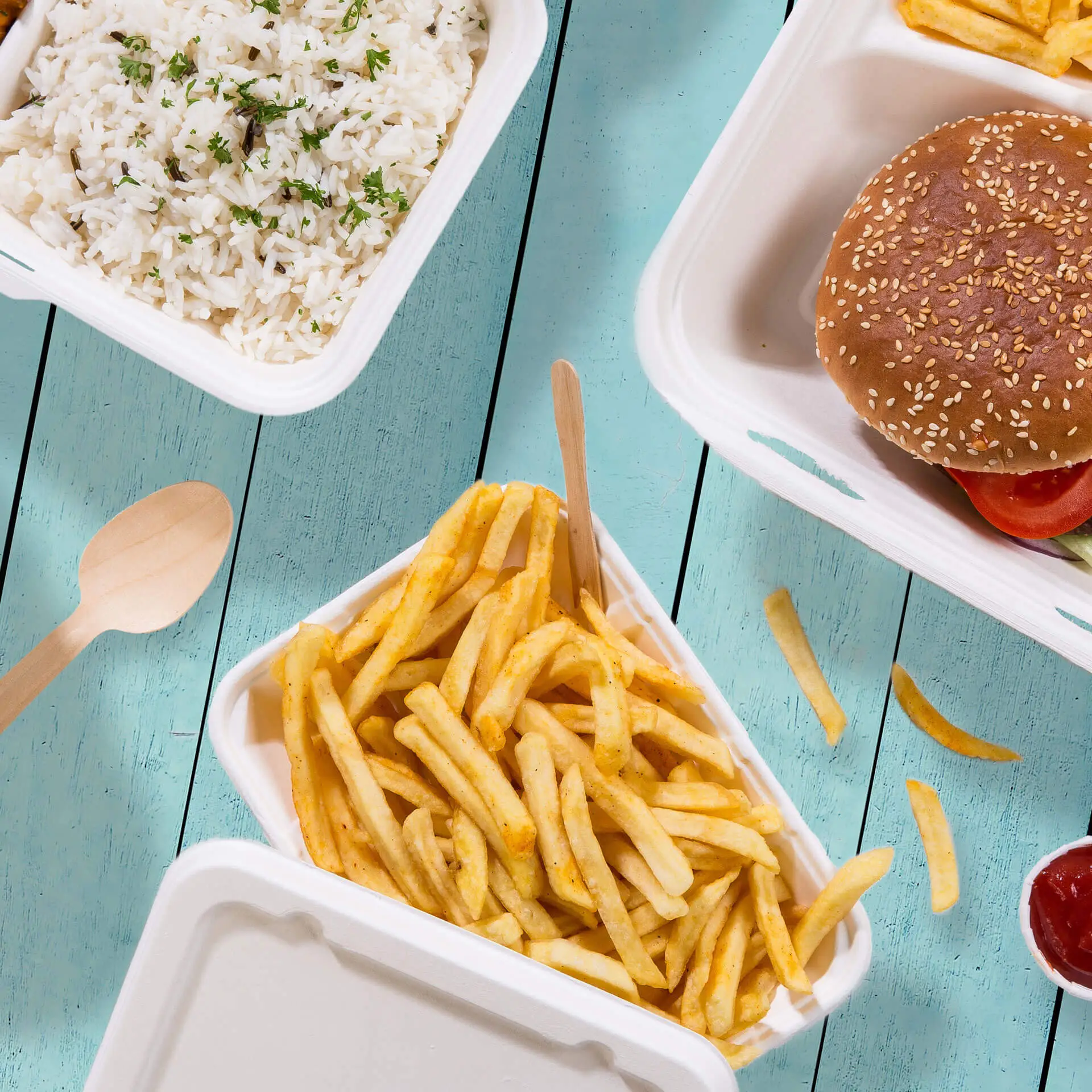 Ciotola biodegradabile in canna da zucchero piena di patatine fritte, cucchiaio di legno accanto; riso e hamburger in contenitori da asporto.
