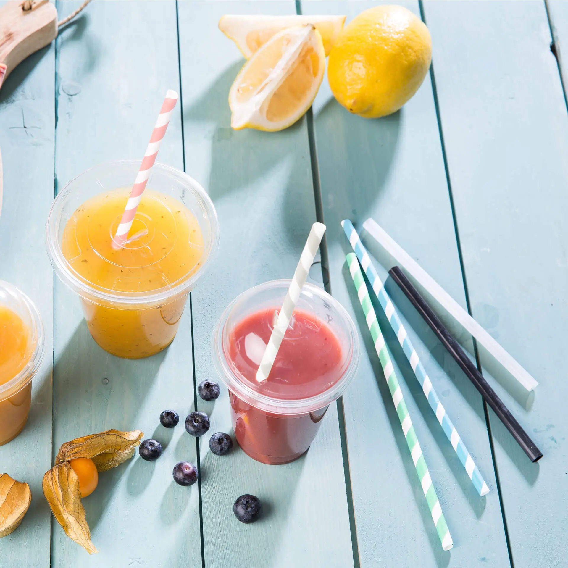 Due bicchieri pieni di bevande arancioni e rosse con cannucce di carta a righe; cannucce pastello e fette di limone fresco nelle vicinanze.