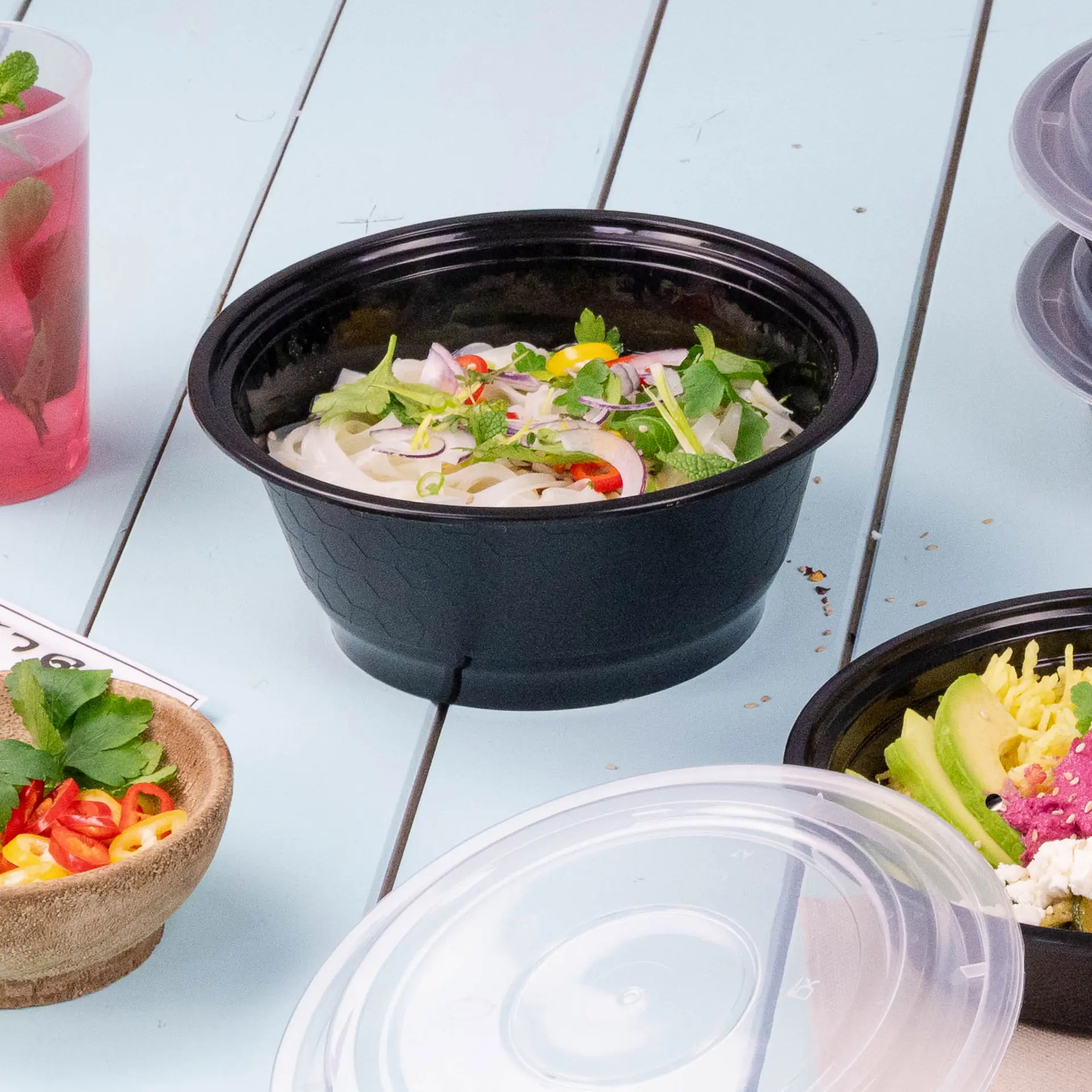 Un contenitore per alimenti riutilizzabile nero pieno di noodles, guarnito con erbe e peperoncini, circondato da altri alimenti.