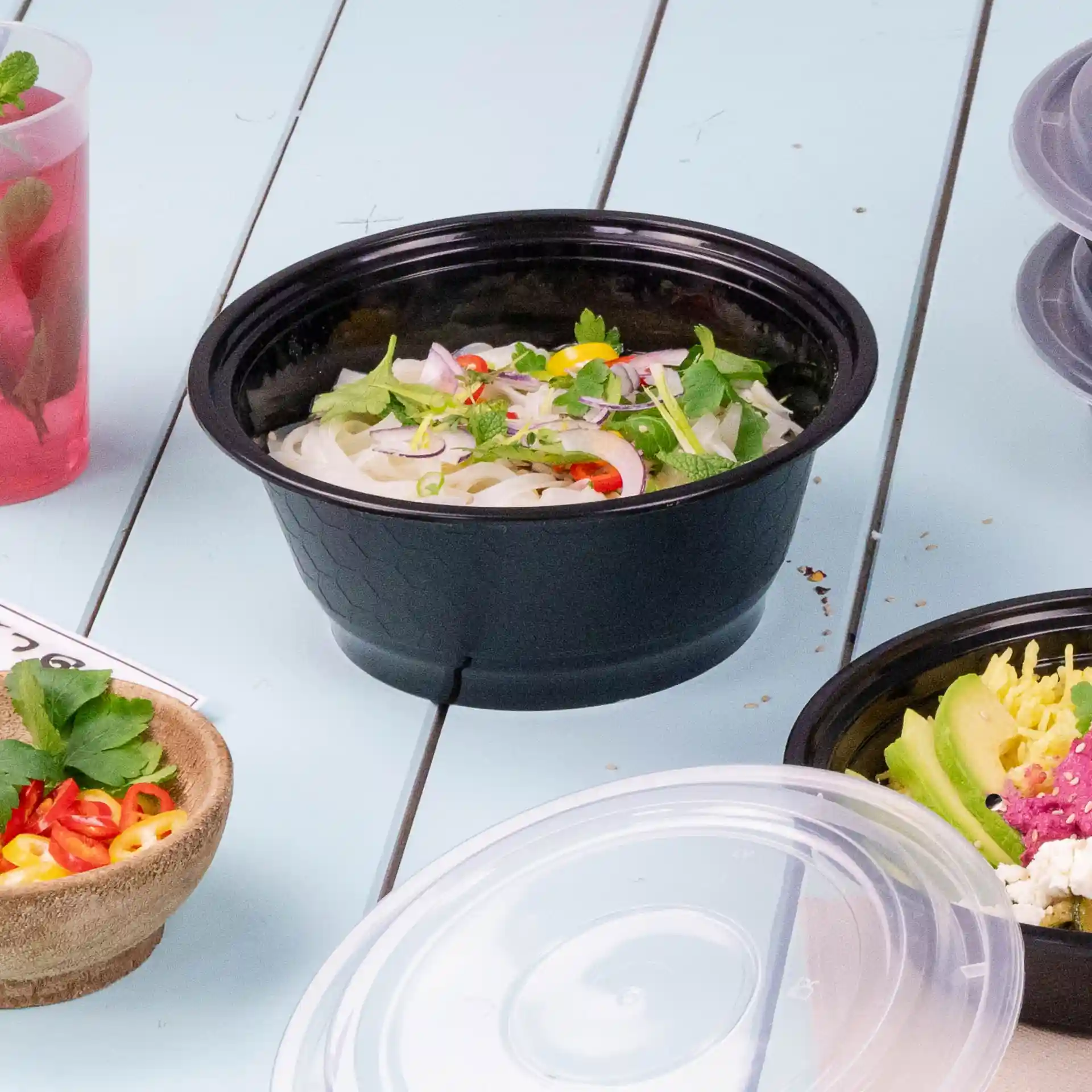 Contenitore riutilizzabile nero riempito di noodles, guarnito con erbe e verdure, su una superficie azzurro chiaro.