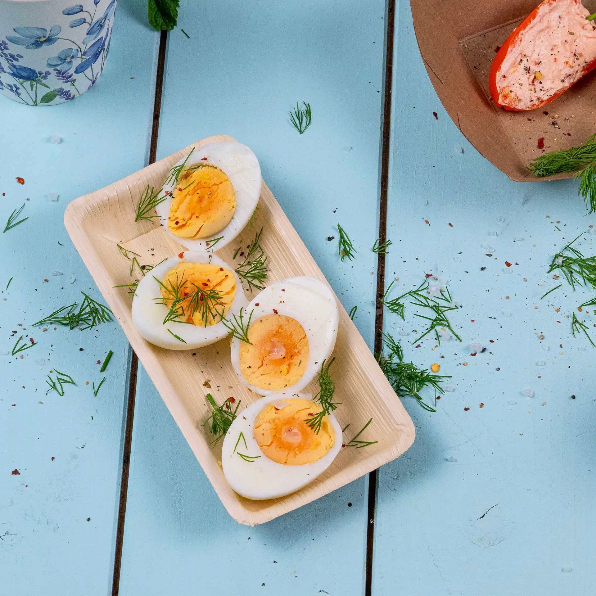 Quattro uova sode tagliate a metà, guarnite con aneto, in una ciotola rettangolare di foglia di palma, con erbe sparse su un tavolo blu chiaro.