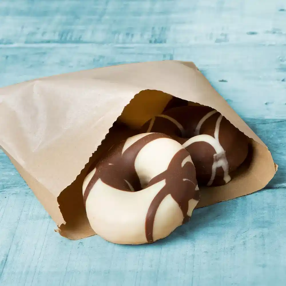 Donuts ricoperti di cioccolato parzialmente visibili in una busta di carta marrone su una superficie in legno blu.