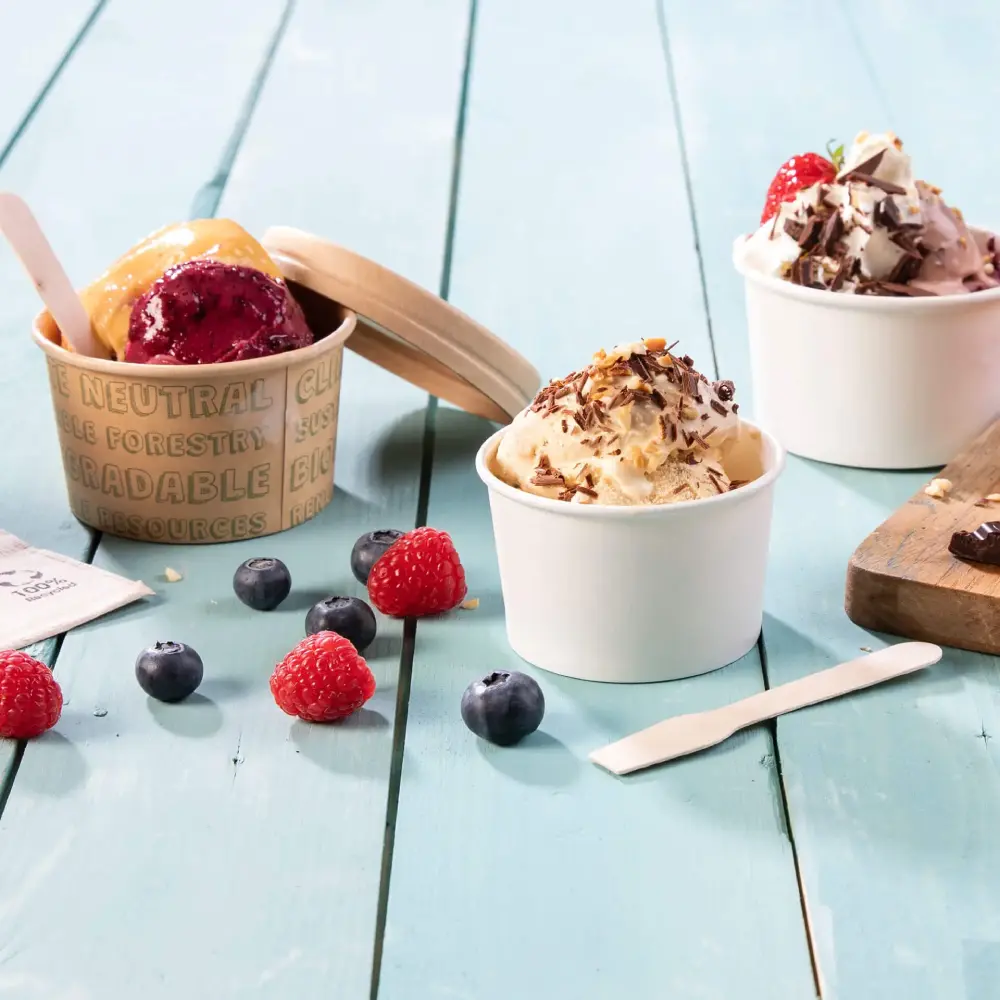 Coppette di gelato con diversi gusti, lamponi freschi, mirtilli e una spatola di legno su una superficie di legno blu.