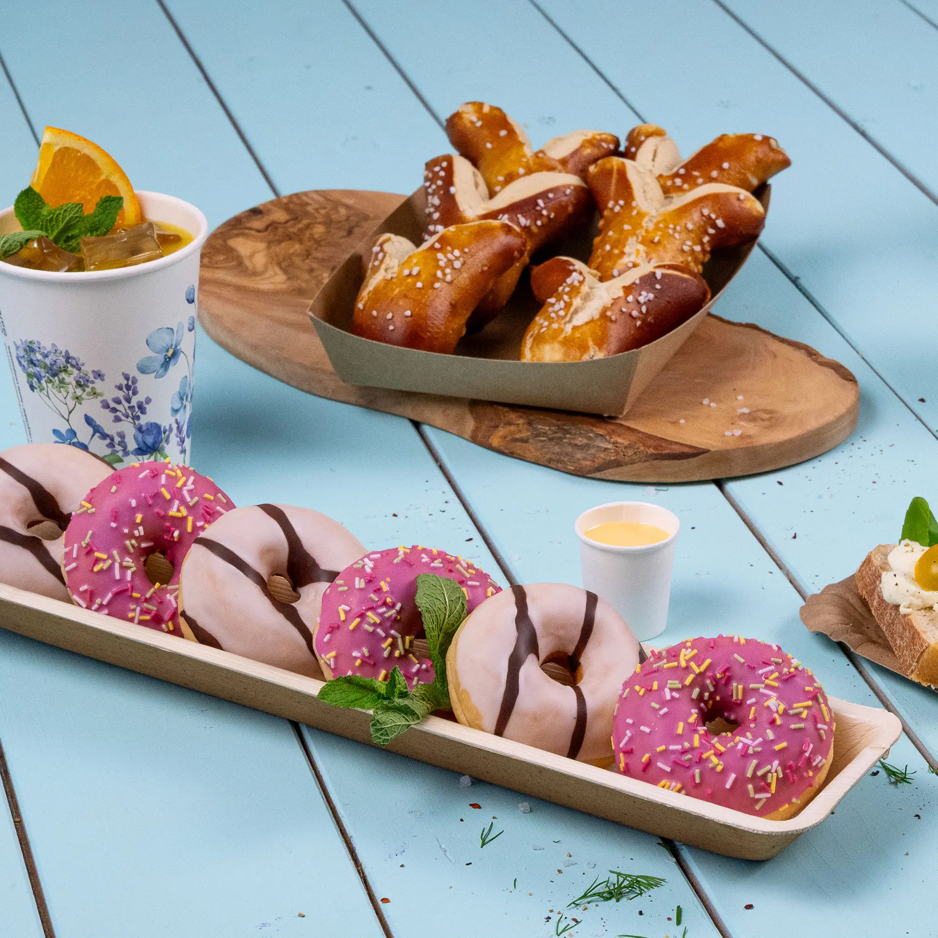 Un vassoio di ciambelle rosa e bianche con glassa al cioccolato, una tazza di bevanda e pretzel su una superficie blu chiaro.