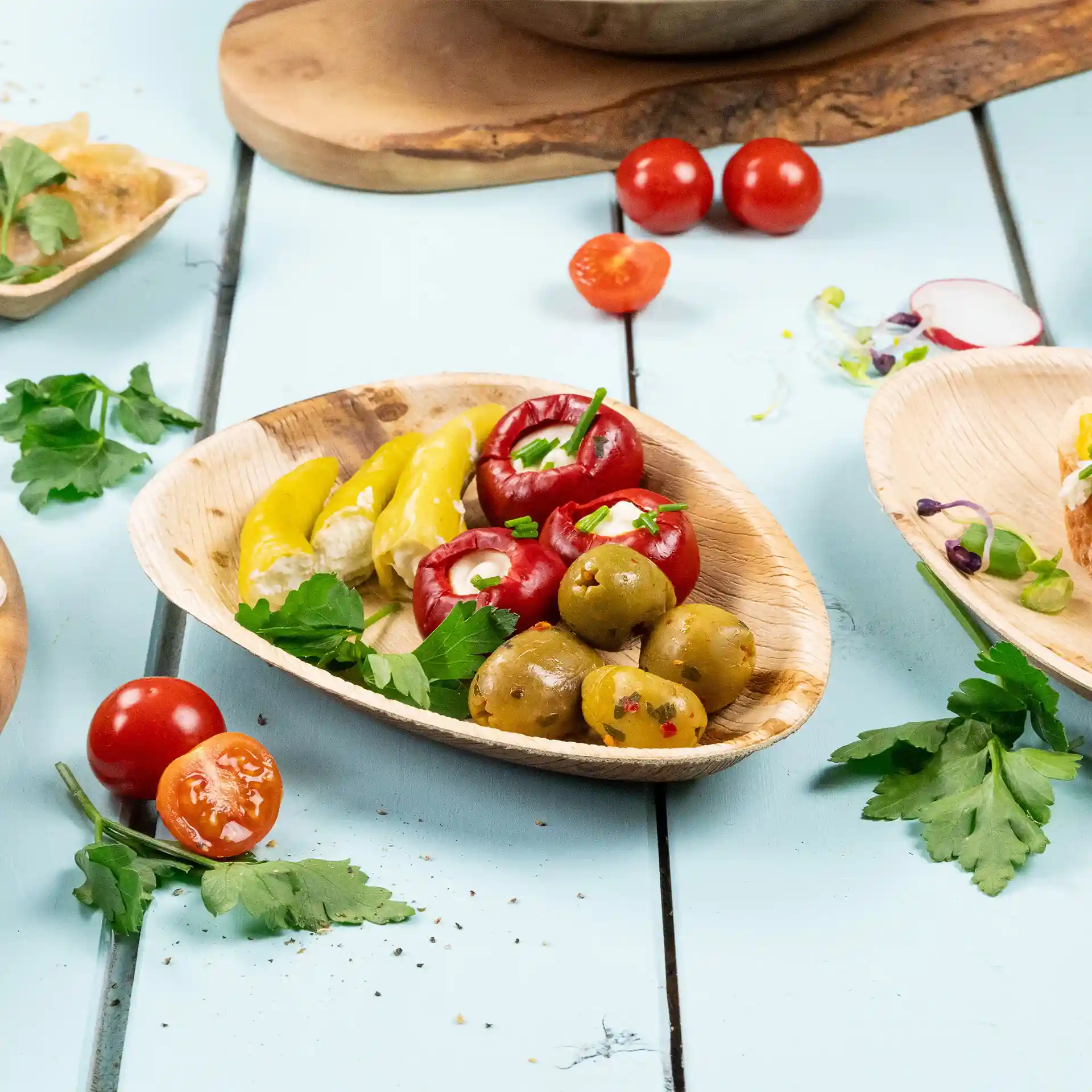 Piatto ovale in foglia di palma con peperoni gialli, pomodori ripieni rossi, olive verdi e prezzemolo su una superficie blu.
