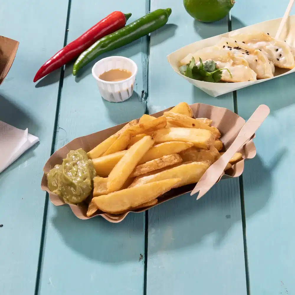 Cestino di patatine fritte con salsa verde, posate di legno e peperoncini su un tavolo di legno blu.