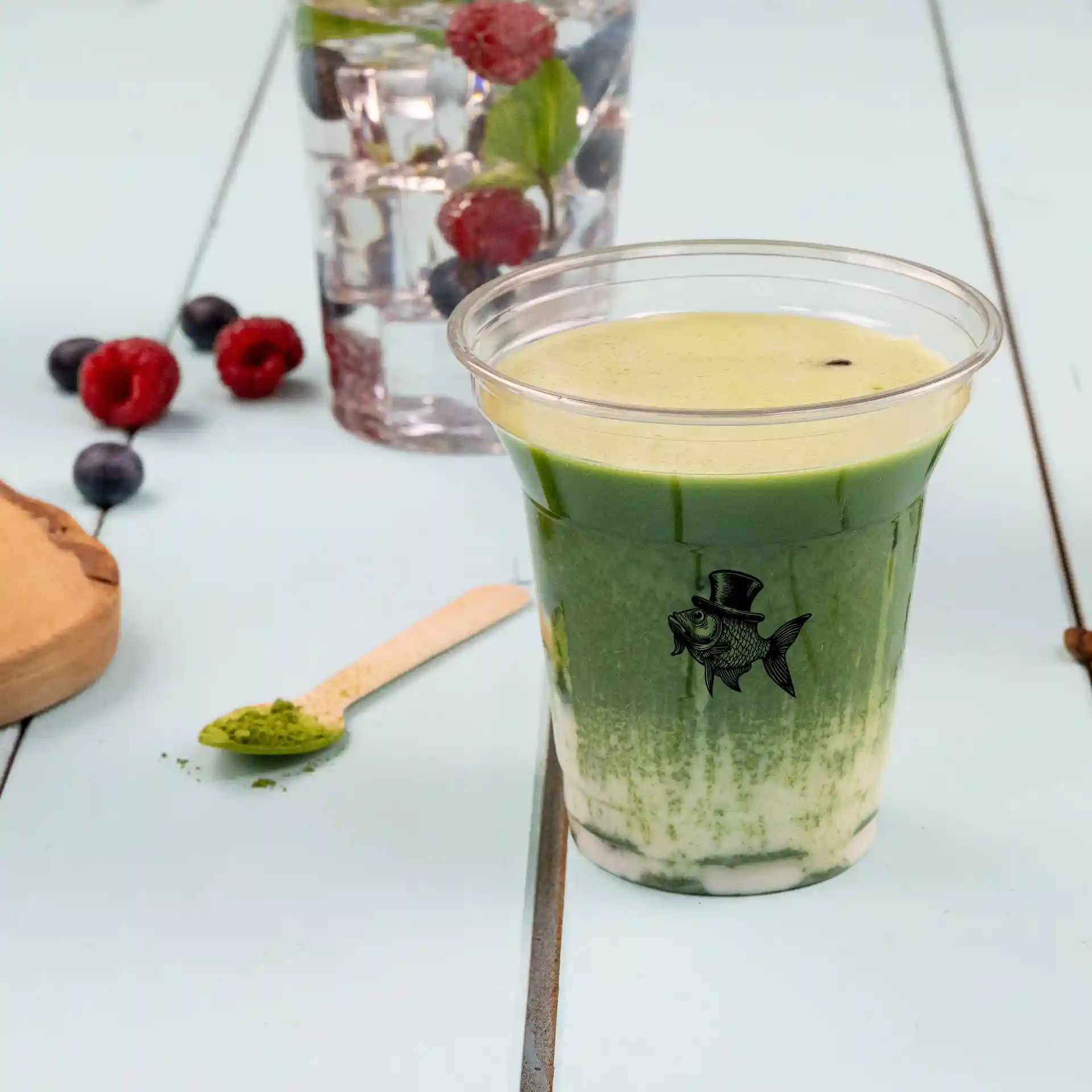 Bicchiere rPET trasparente da 8 oz riempito con bevanda verde, con logo di pesce, cucchiaio di legno e bacche sullo sfondo.
