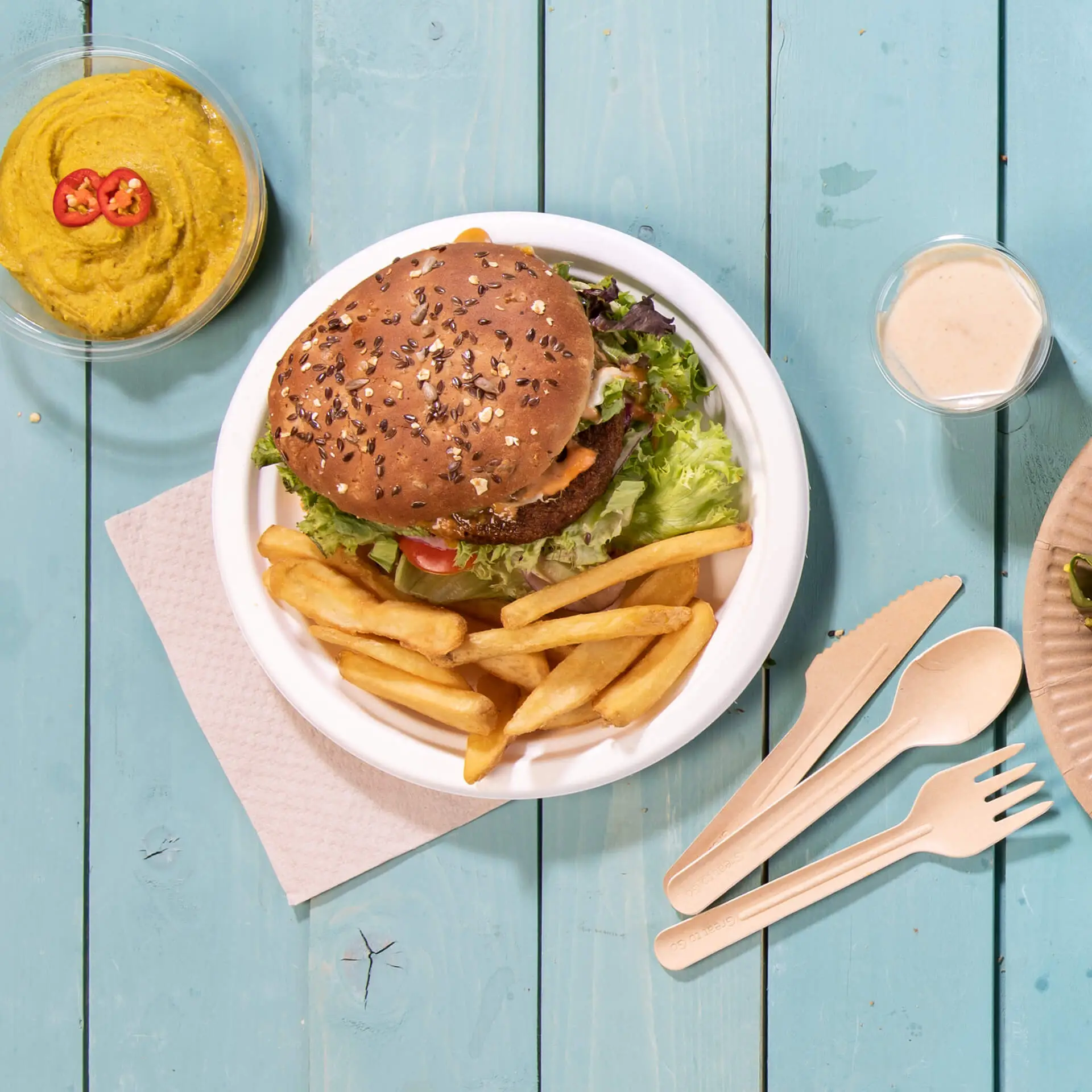 Un piatto di cibo con un hamburger al sesamo marrone, lattuga e patatine, accompagnato da un contenitore di hummus e posate usa e getta.