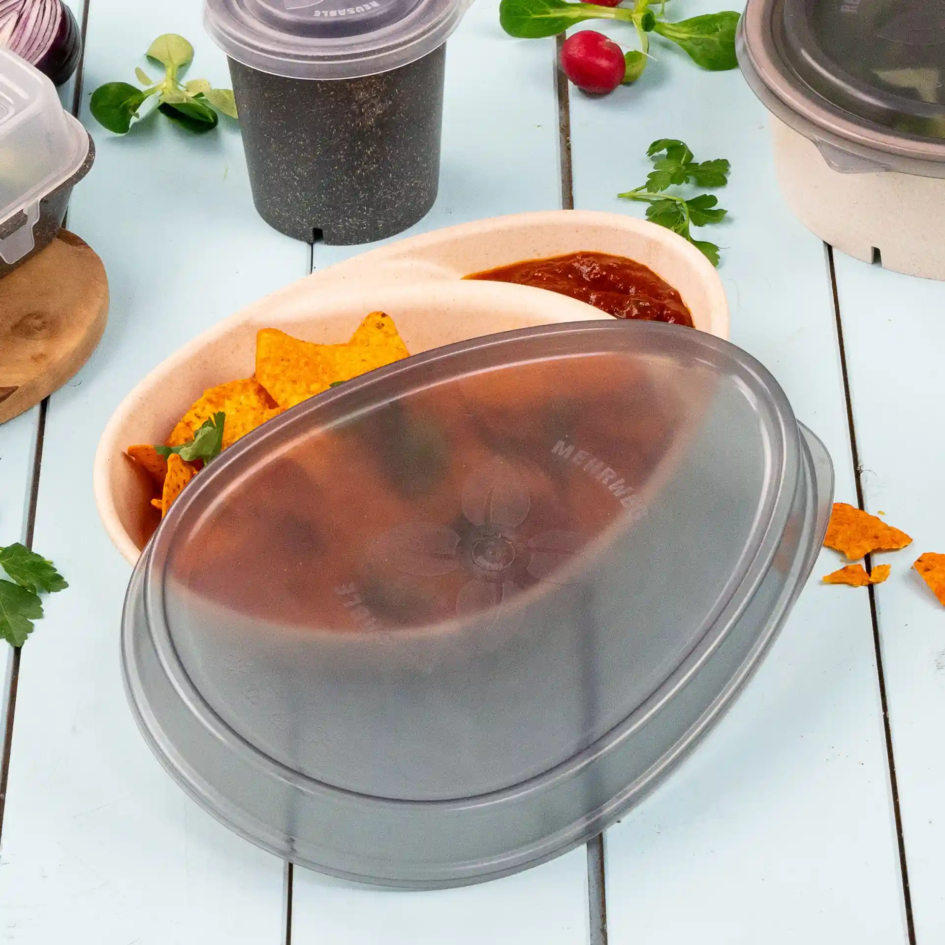 Coperchio riutilizzabile grigio ovale per un piatto, mostrato con tortilla chips e salsa in un contenitore, su una superficie azzurro chiaro.
