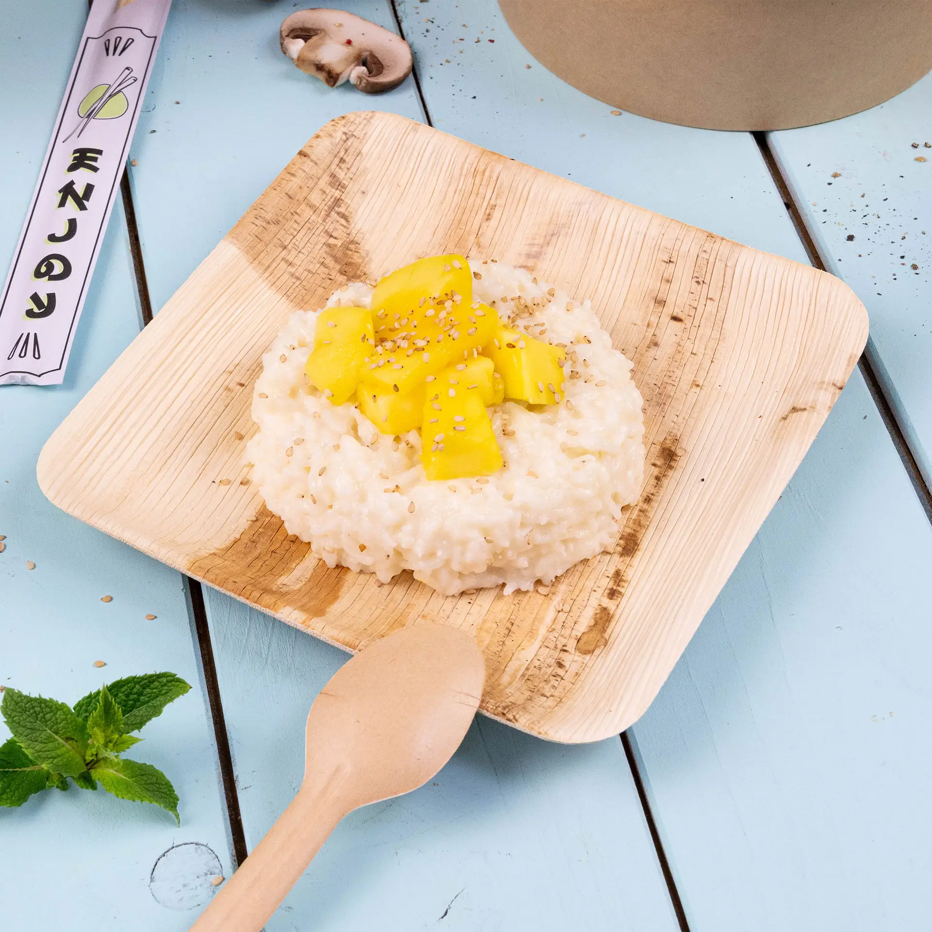 Piatto quadrato in foglia di palma con risotto guarnito con pezzi di mango e semi di sesamo, su uno sfondo blu con un cucchiaio di legno.