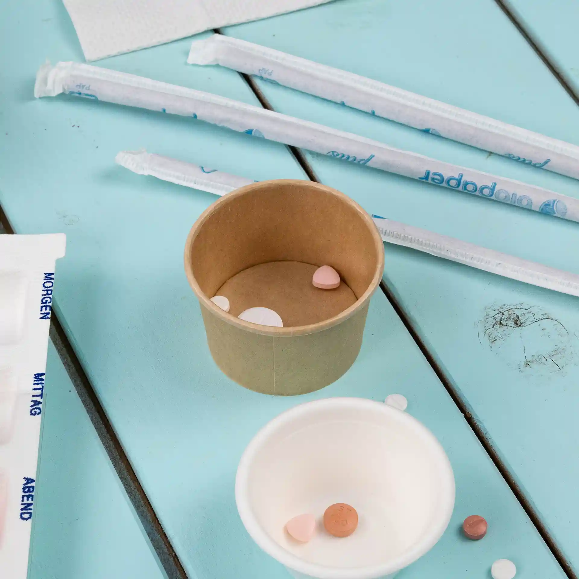 Cup in carta marrone con diverse piccole pillole all'interno, posato su una superficie in legno blu con cannucce vicine.