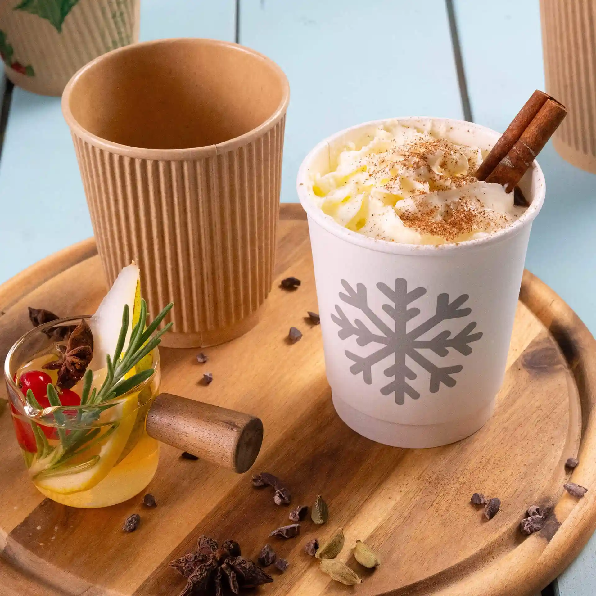 Bicchiere bianco con design di fiocco di neve, riempito di panna montata e un bastoncino di cannella, accanto a un bicchiere kraft e una bevanda guarnita.