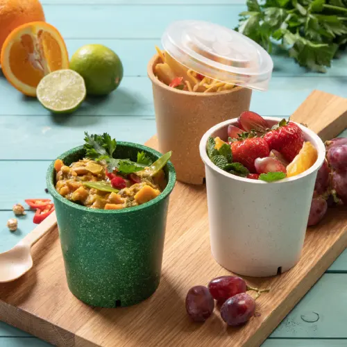 Tre tazze per zuppa riutilizzabili su un tagliere di legno, piene di zuppa, frutta e noodles, circondate da agrumi freschi e uva.