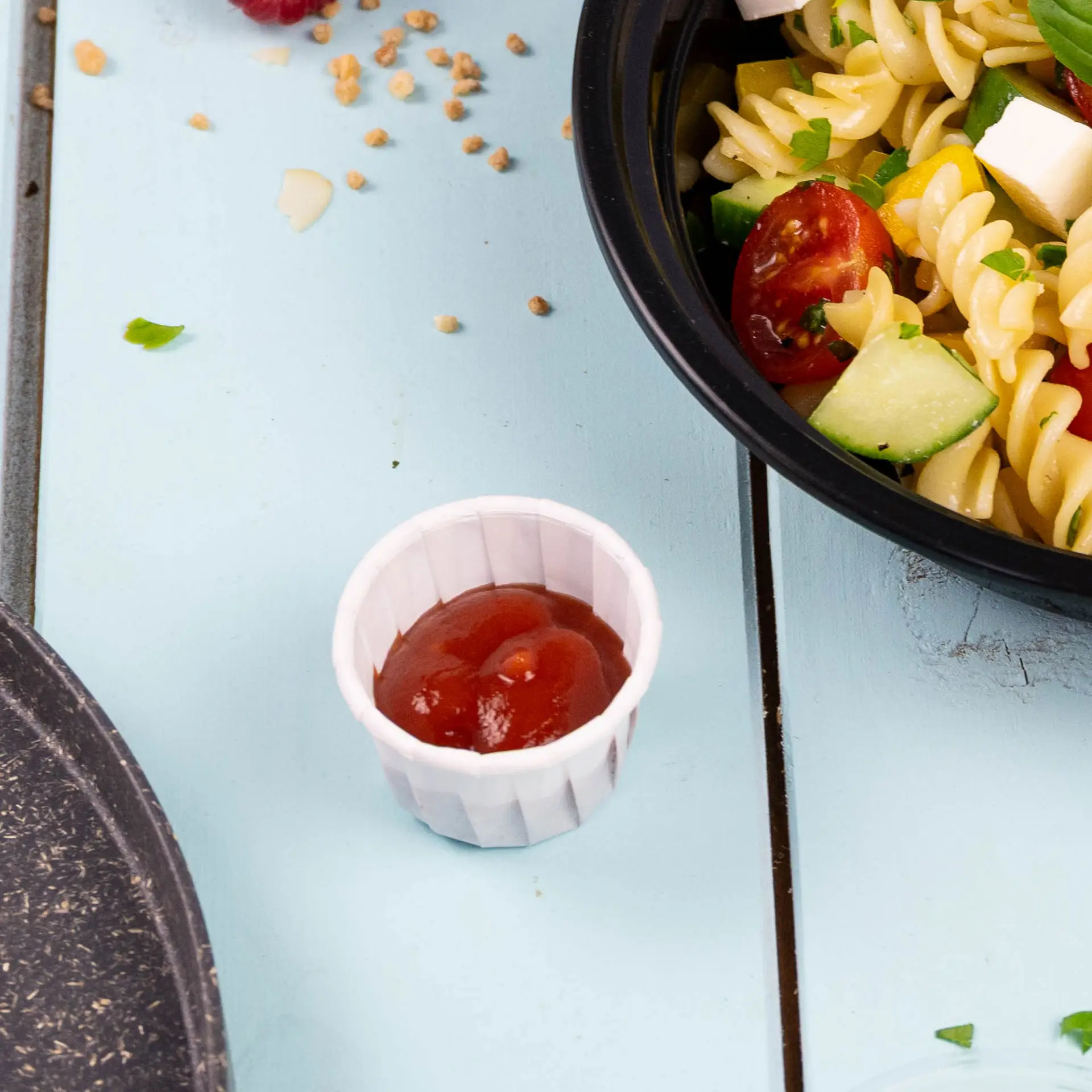 Piccolo bicchiere di carta bianco pieno di ketchup, posizionato accanto a una ciotola di insalata di pasta su una superficie di legno blu.